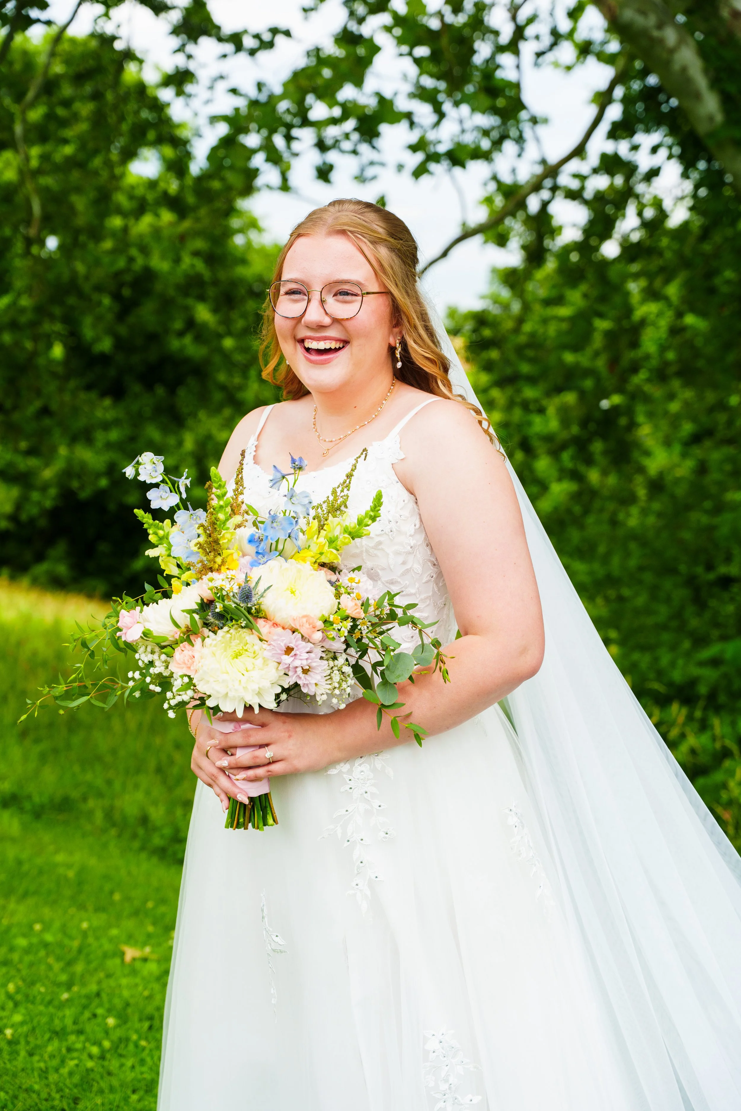 A smiling bride in a white wedding dress holding a bouquet of flowers outdoors with lush green trees in the background.