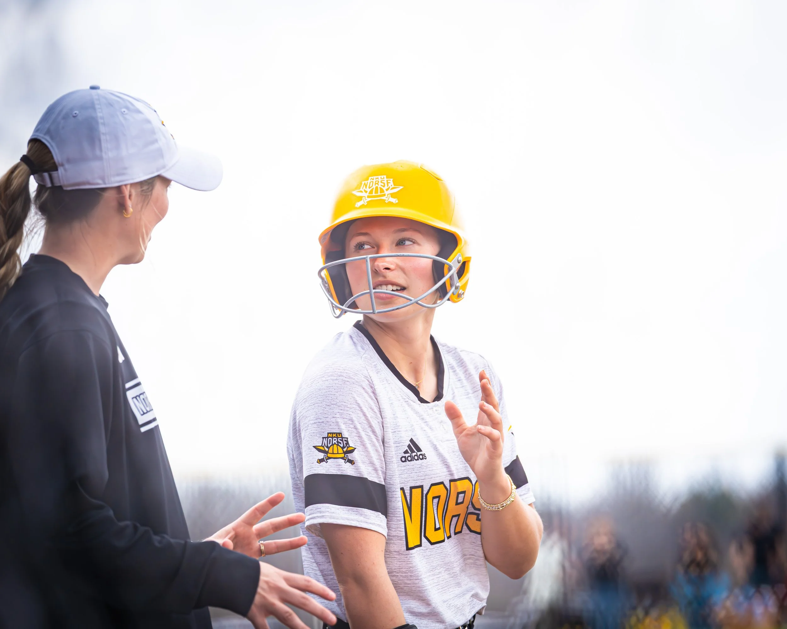 Two women, one in a white team jersey and helmet, and the other in a black jacket and white baseball cap, are engaged in a conversation outdoors, with the woman in the helmet smiling and gesturing with her hands.