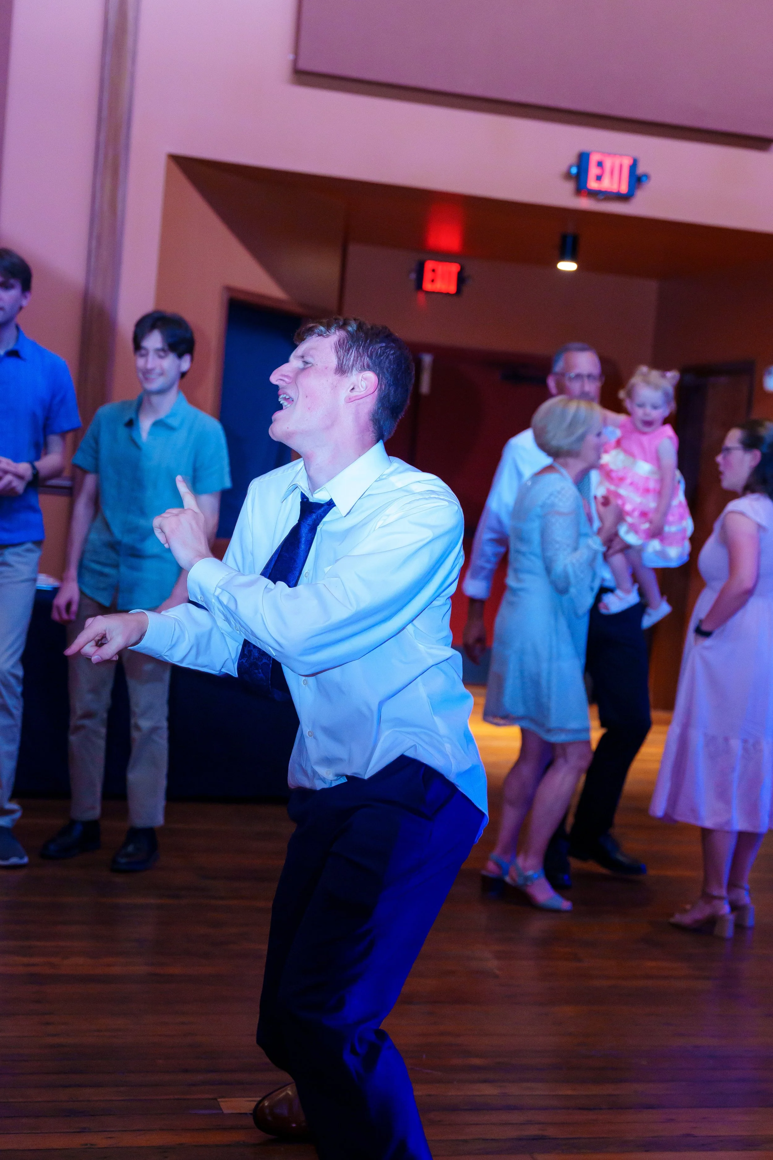 People dancing at a social event, with a man in a white shirt and tie in the foreground, smiling and gesturing, and a group of men, women, and children in the background.