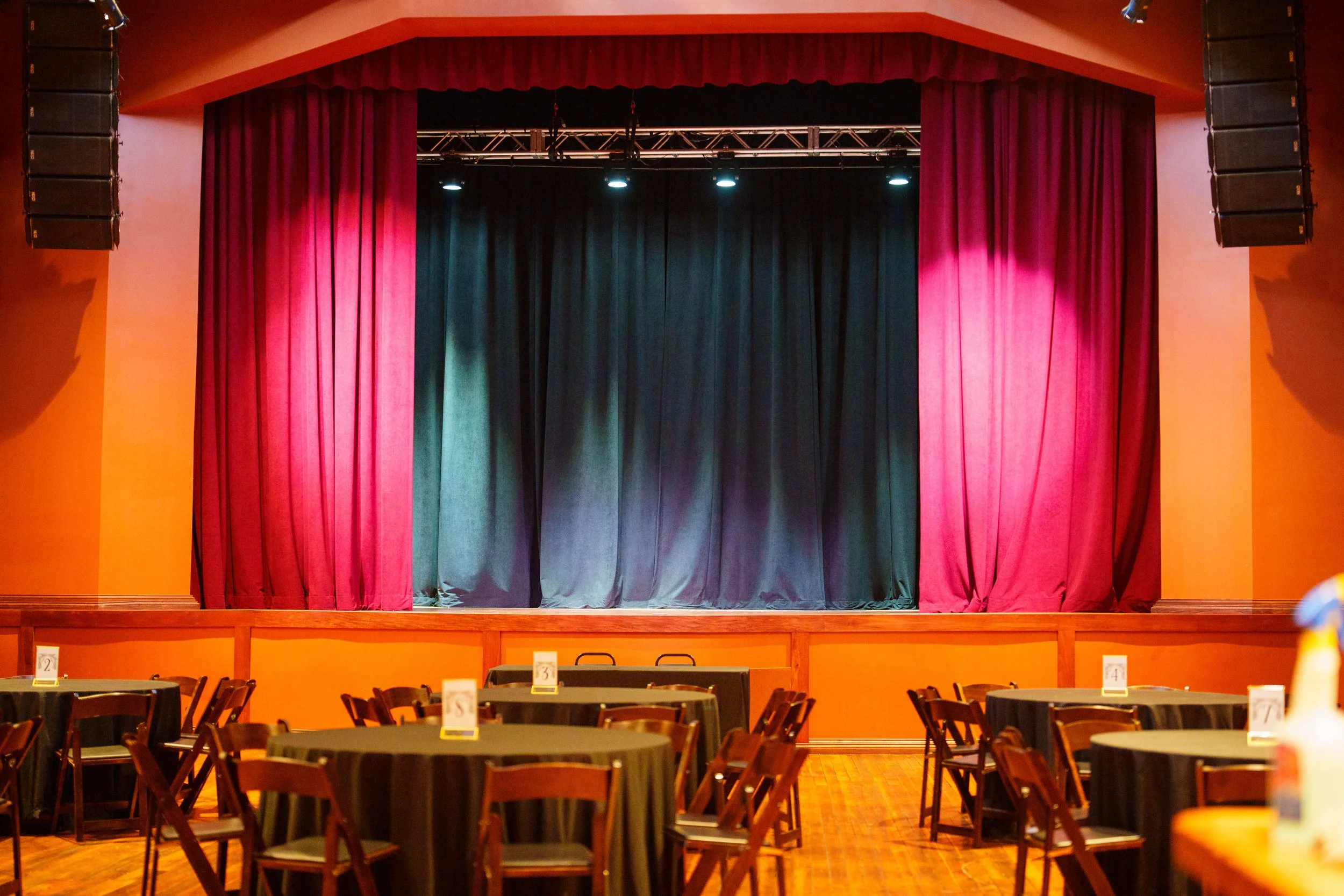 A stage with closed red curtains in a theater or event hall, with round tables and chairs set up in front for an audience or guests.