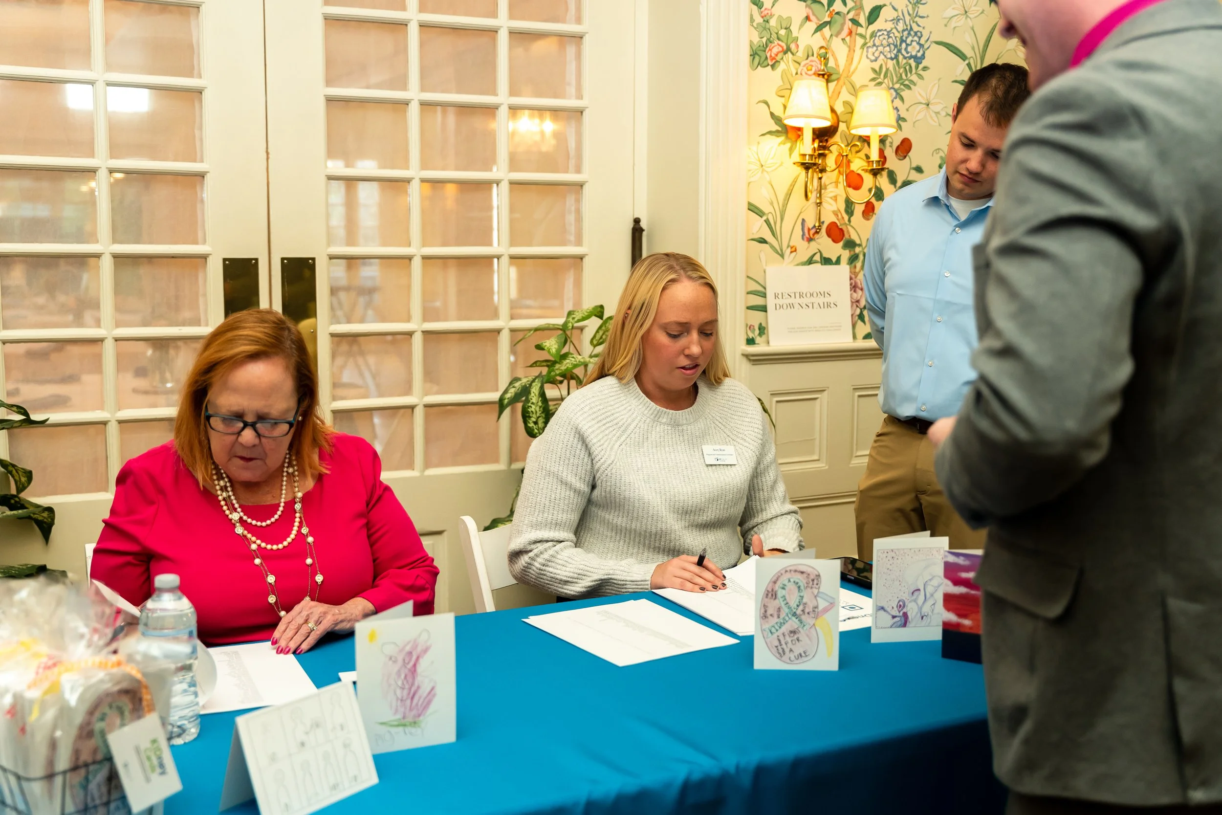 People sitting at a table with artwork and pamphlets, engaging in what appears to be a book signing event or fundraiser in an elegant room with floral wallpaper and wall sconces.