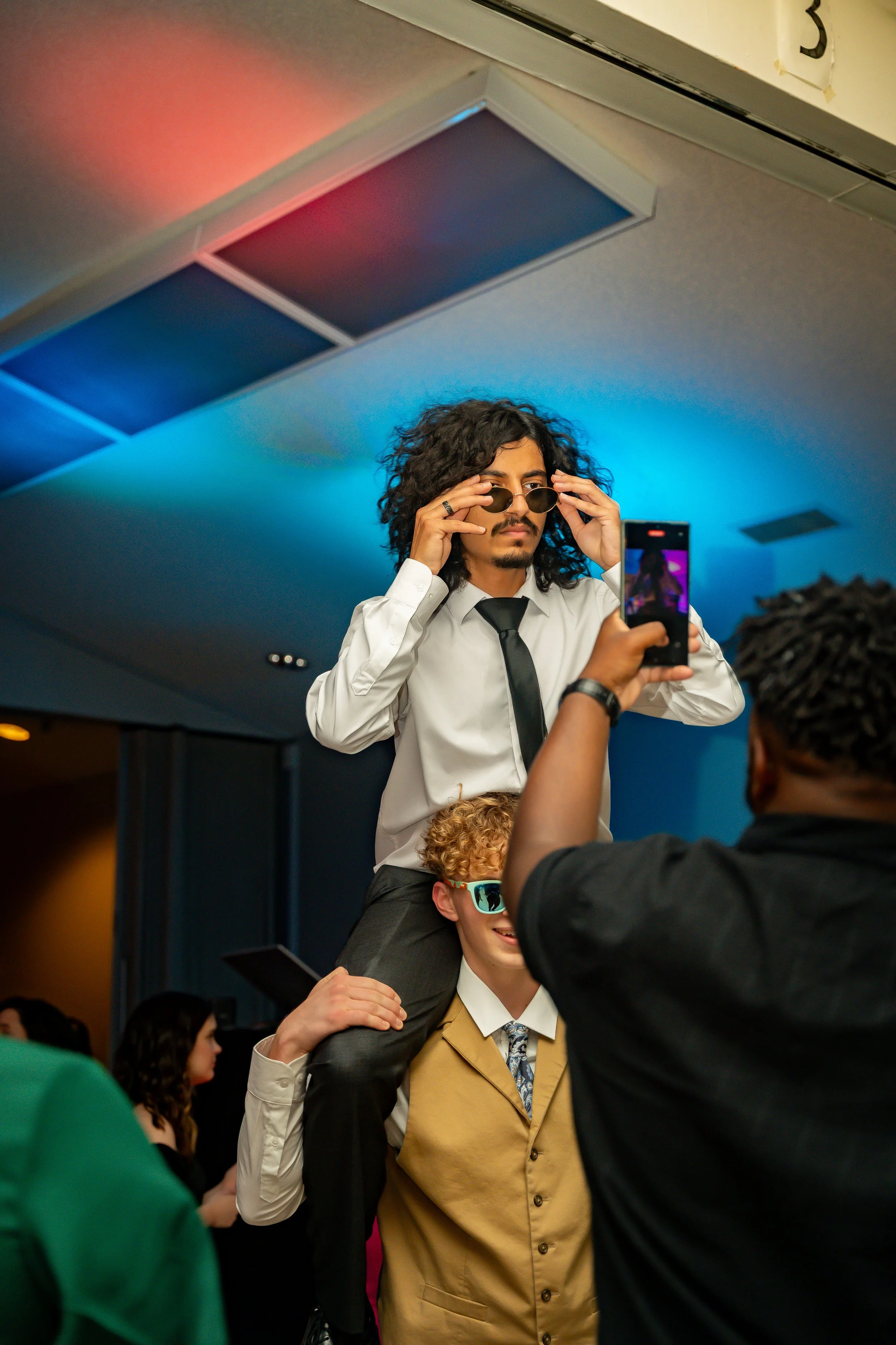 A man with curly hair, in a white shirt and black tie, is sitting on another person's shoulders. He is adjusting his sunglasses as others are around him, including a person taking a photo and a young boy. The scene appears to be at an indoor event or