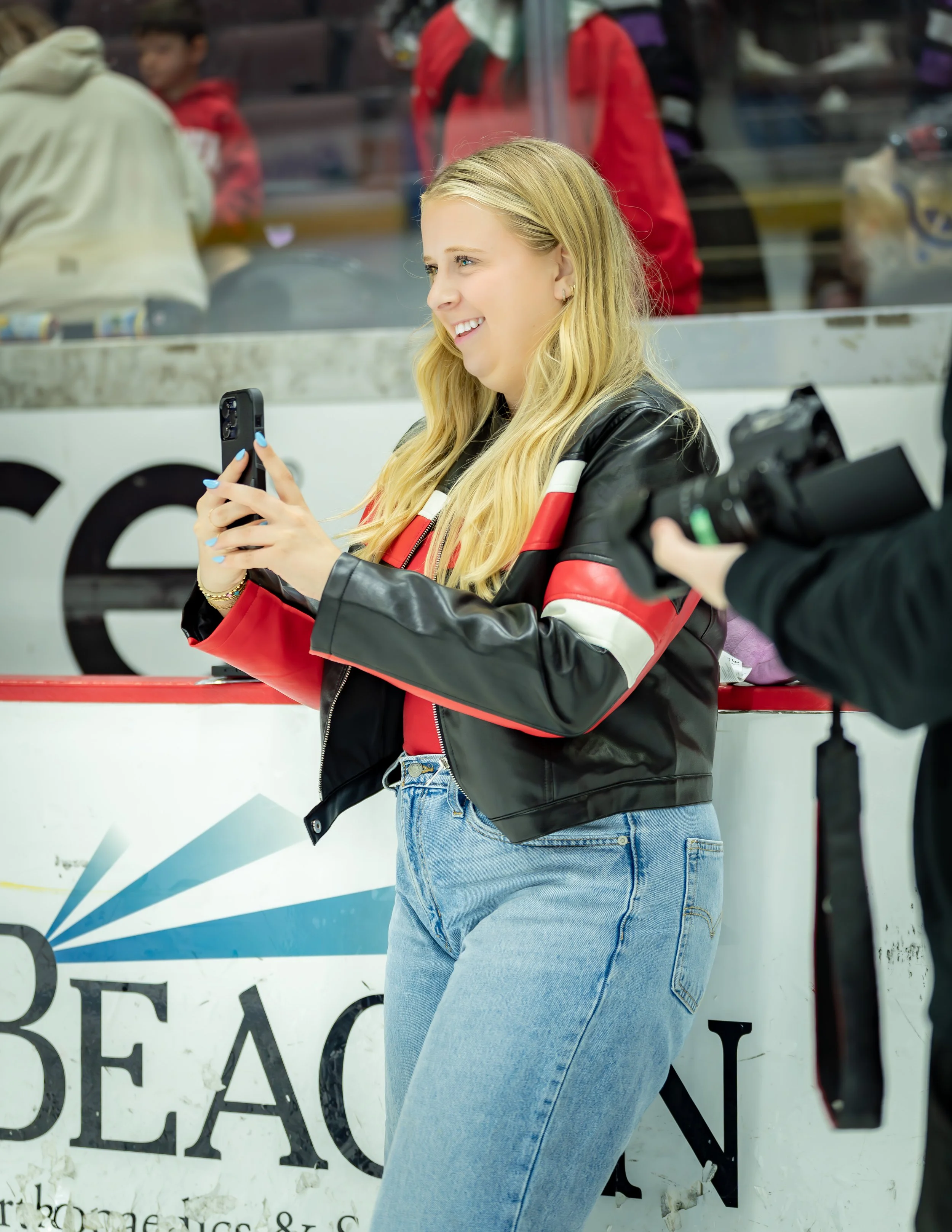 A young woman with long blonde hair smiling, wearing a black and red jacket and light blue jeans, standing in front of an ice skating rink, holding a phone in her hand.