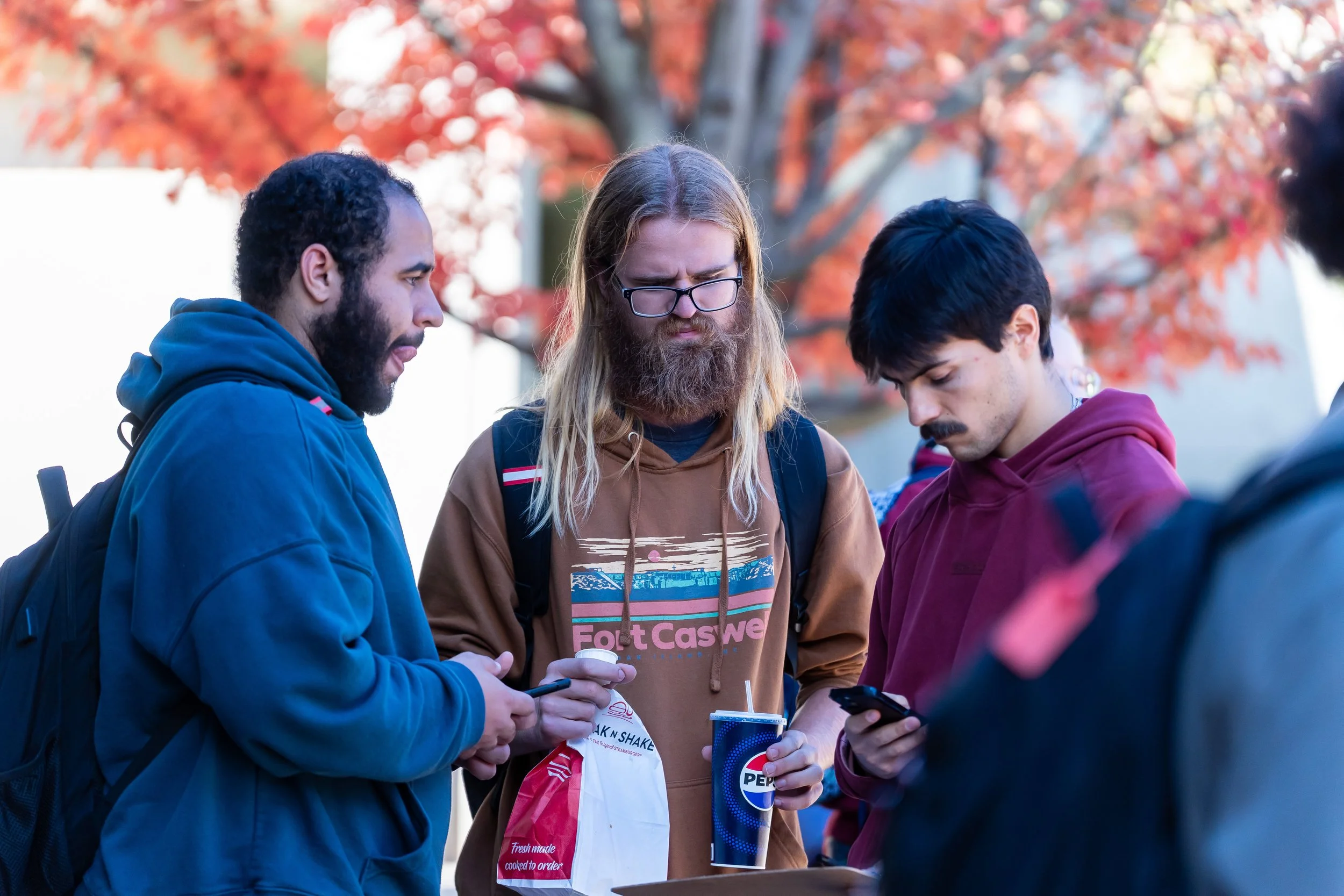 Three young men are standing outdoors in front of a tree with red leaves, looking at their smartphones. They are dressed casually, and there are drinks and a food bag in their hands.