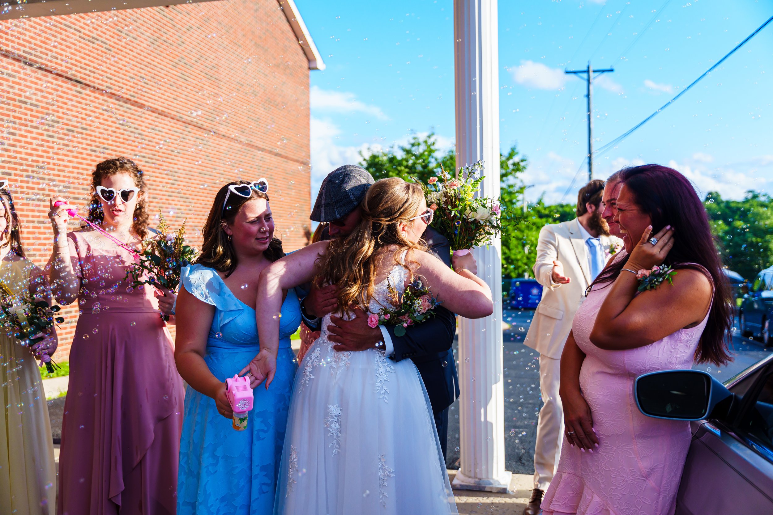A group of women and men celebrating at a wedding outdoors, with some women wearing sunglasses and holding flowers, and bubbles floating in the air.