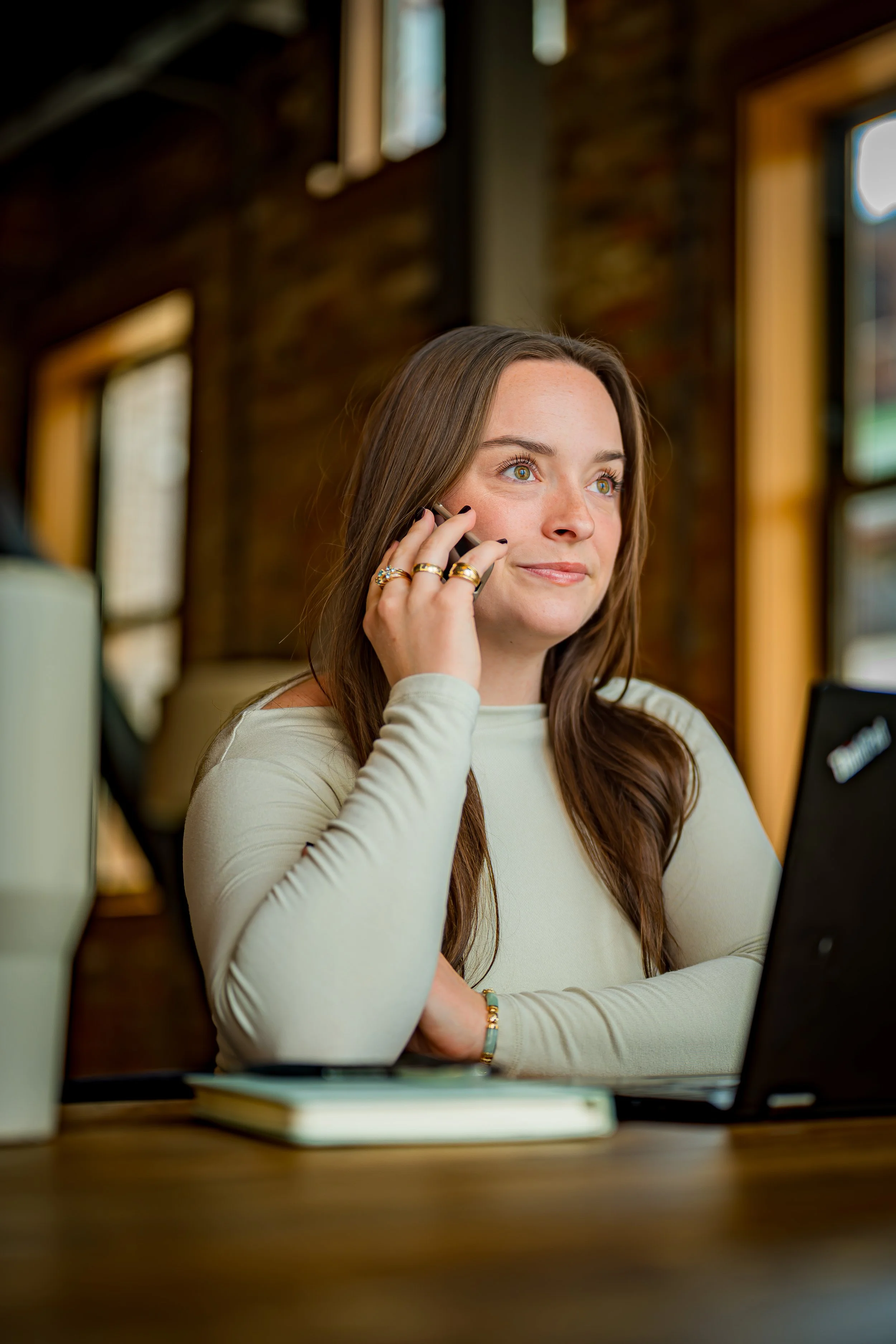 A young woman with long brown hair sitting at a desk in a cozy, rustic-style room with exposed brick walls. She is smiling slightly, holding a phone to her ear, and looking slightly to the side. There is a closed laptop, a notebook, and some papers on the desk in front of her.