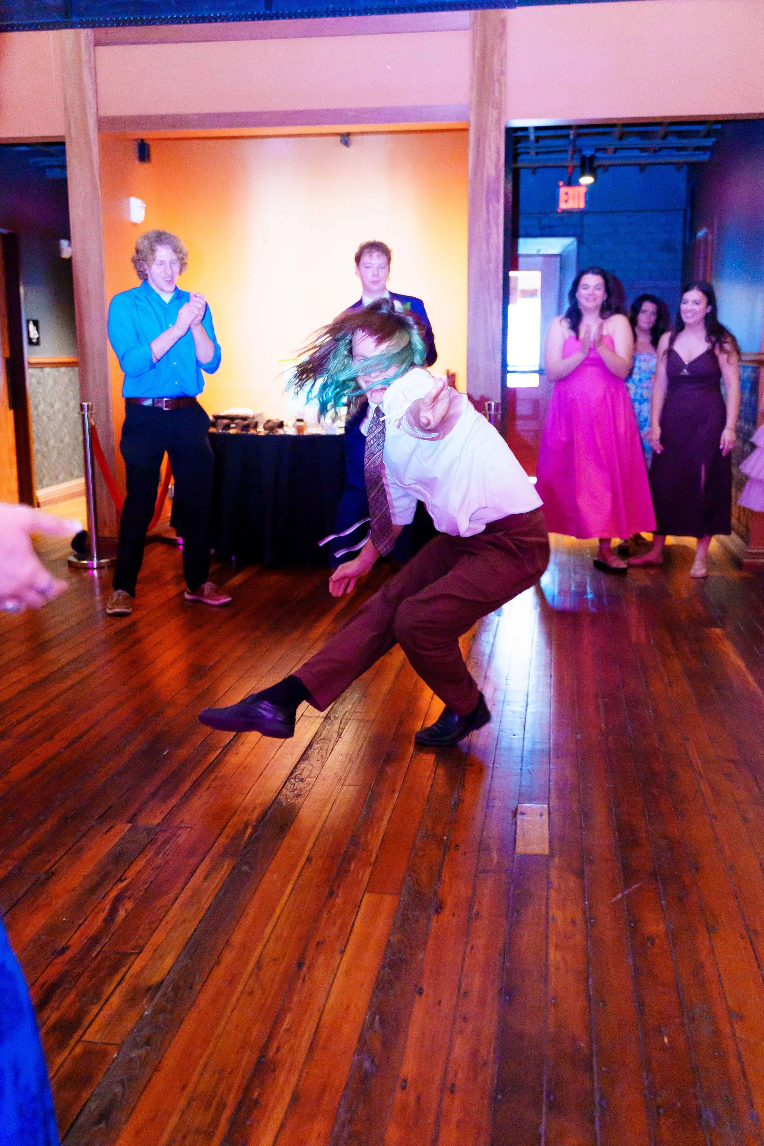 A person with colorful hair, wearing a white shirt and brown pants, is dancing or performing dance moves on a wooden floor in front of a group of people. The group is clapping and smiling, watching the performance. The setting appears to be an indoor