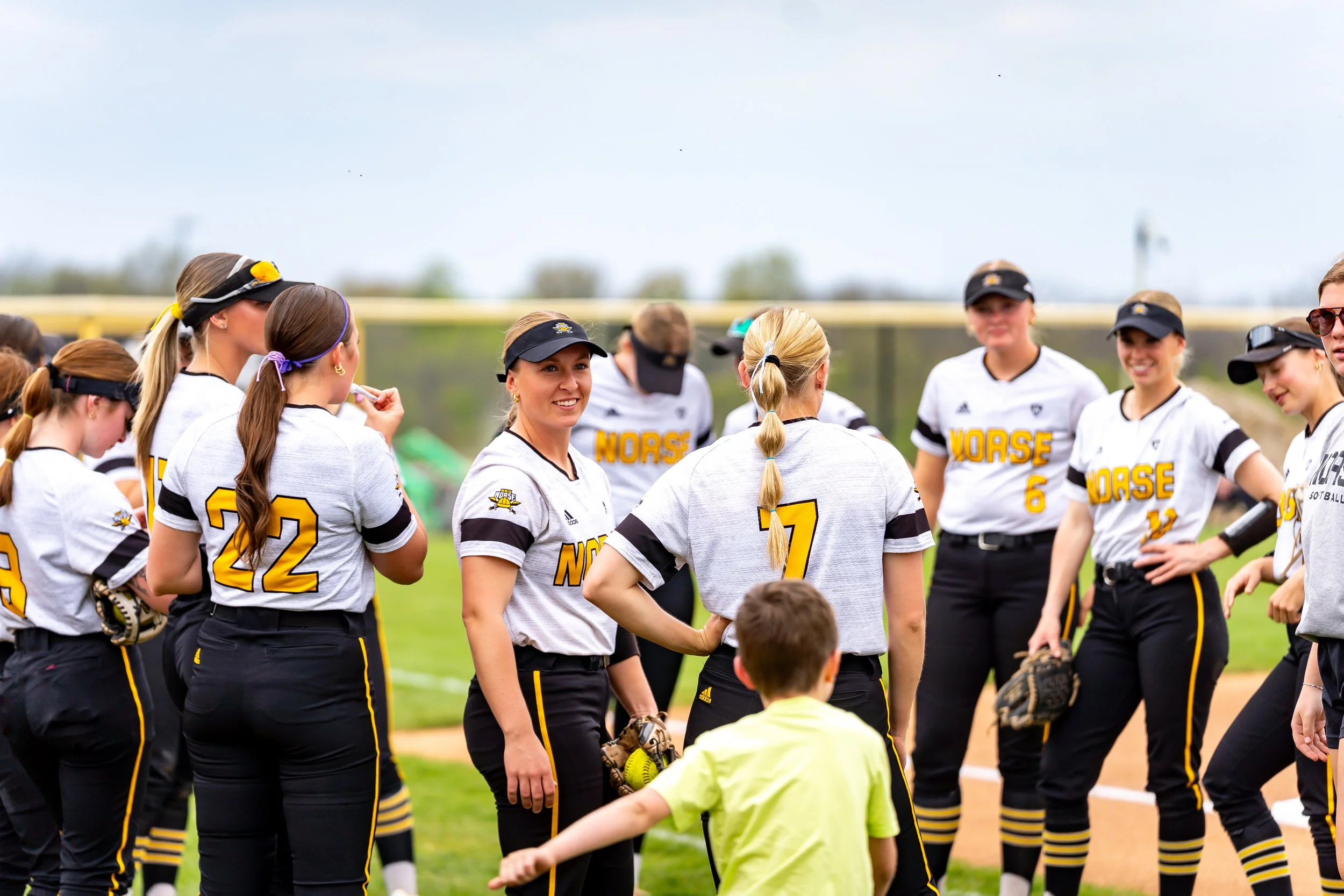 Women softball players in black and yellow uniforms standing on a field, some wearing caps, with a few children nearby, under a cloudy sky.