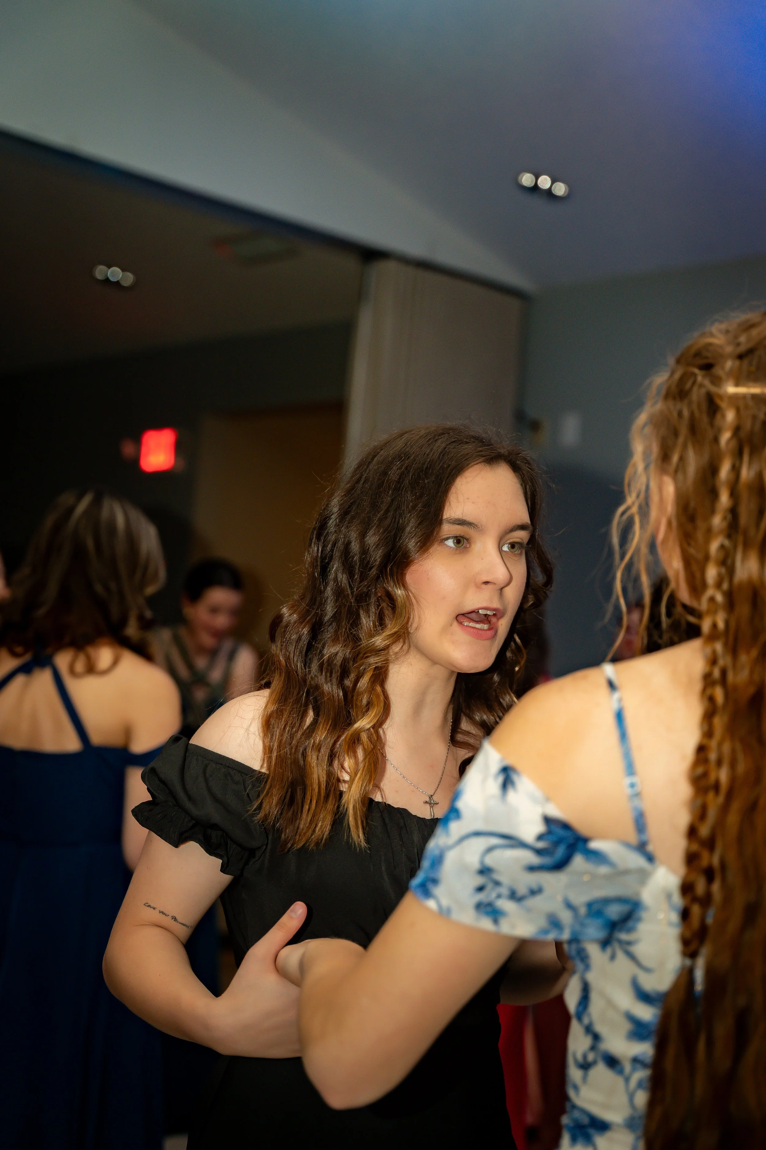 Young woman with long wavy brown hair, wearing a black dress with off-the-shoulder ruffled sleeves, talking to another woman with curly red hair and glasses, in a crowded indoor setting.