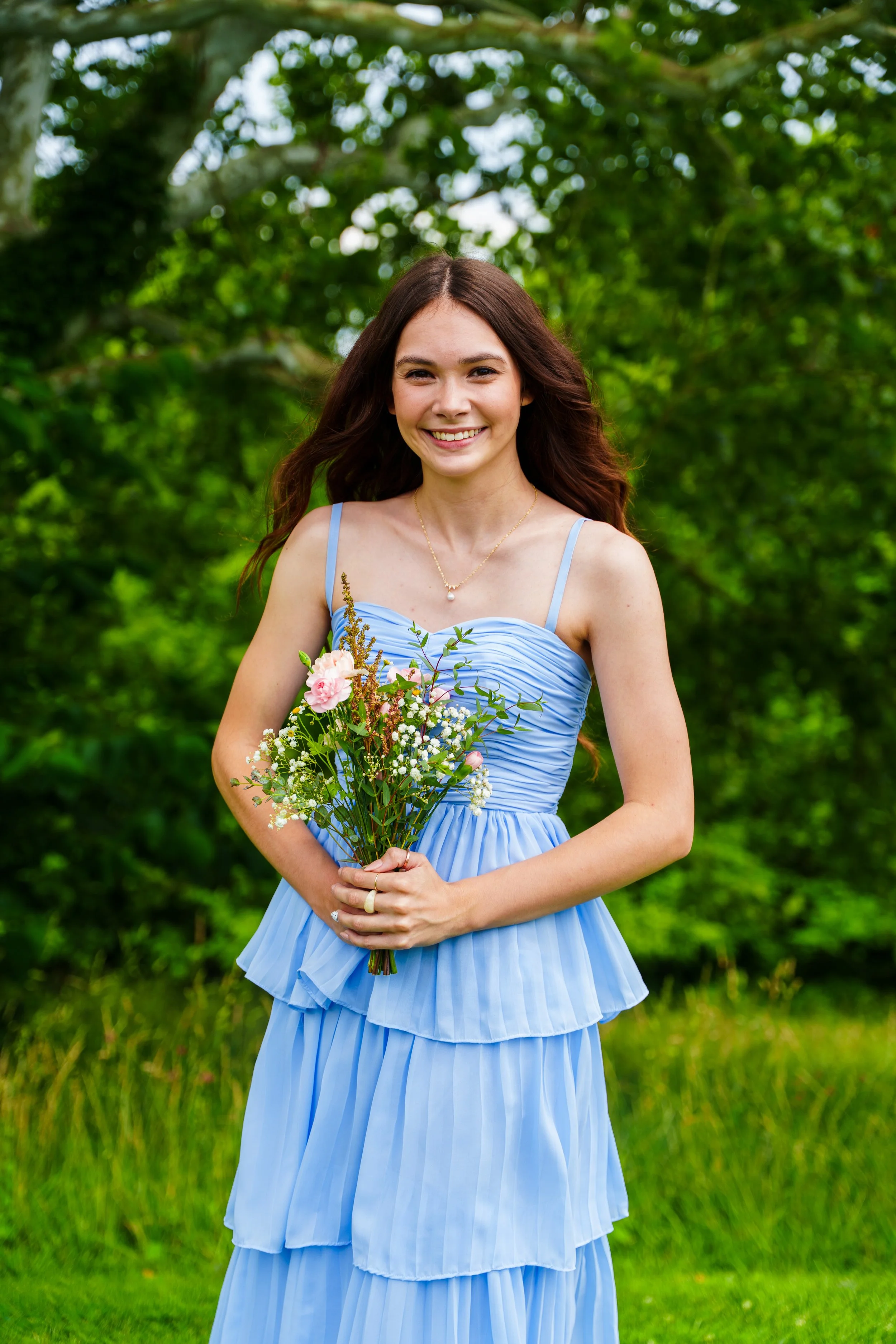 A young woman with long brown hair, smiling, wearing a light blue dress with spaghetti straps and holding a bouquet of pink and white flowers in front of a lush green garden.