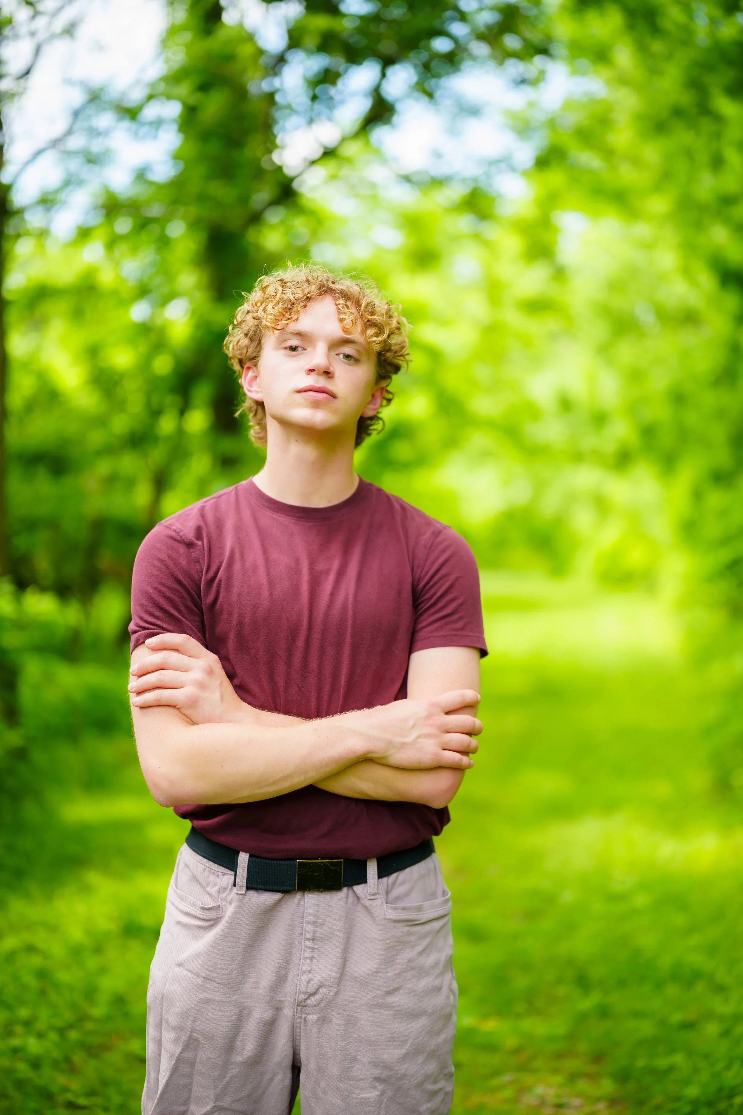 A young man with curly blonde hair wearing a maroon T-shirt and beige pants with a black belt, standing outdoors against a background of green trees and grass, with his arms crossed.