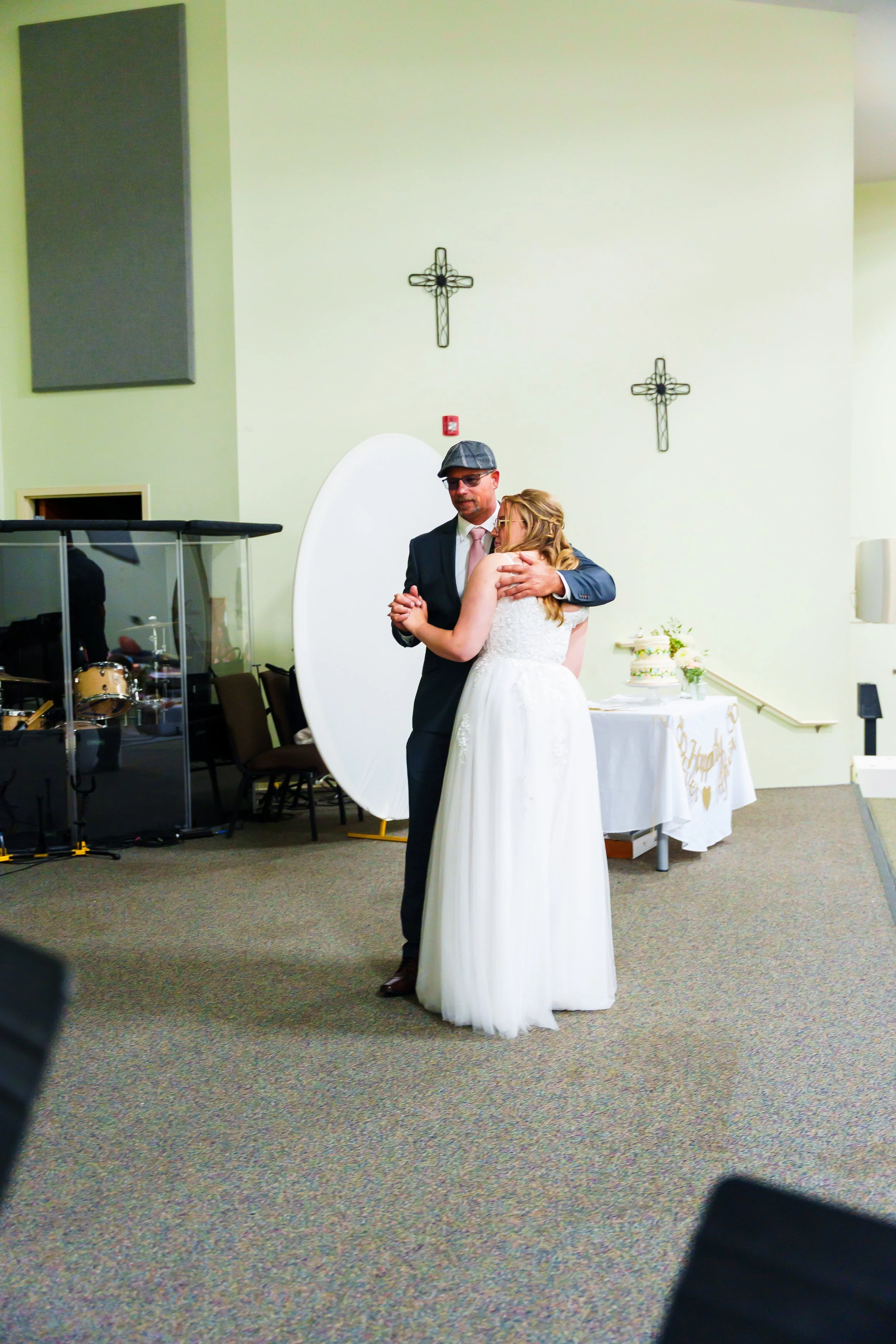 A bride and groom are dancing closely during a wedding reception in a church or community hall with pale green walls and religious crosses on the wall. The groom is wearing a suit and a flat cap, and the bride is in a white wedding dress. A wedding c