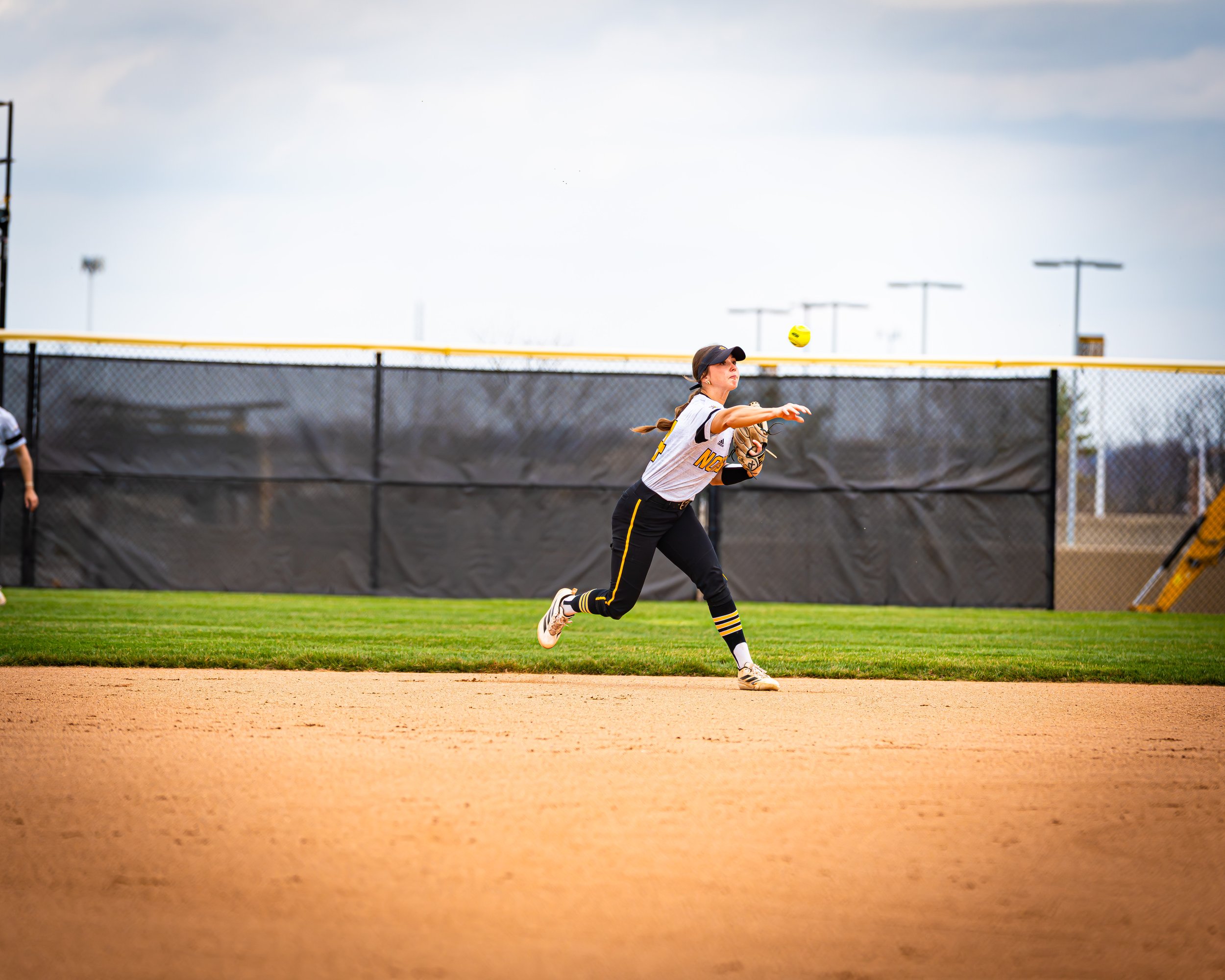 A young girl in sports uniform playing softball on a field, preparing to catch a yellow ball.