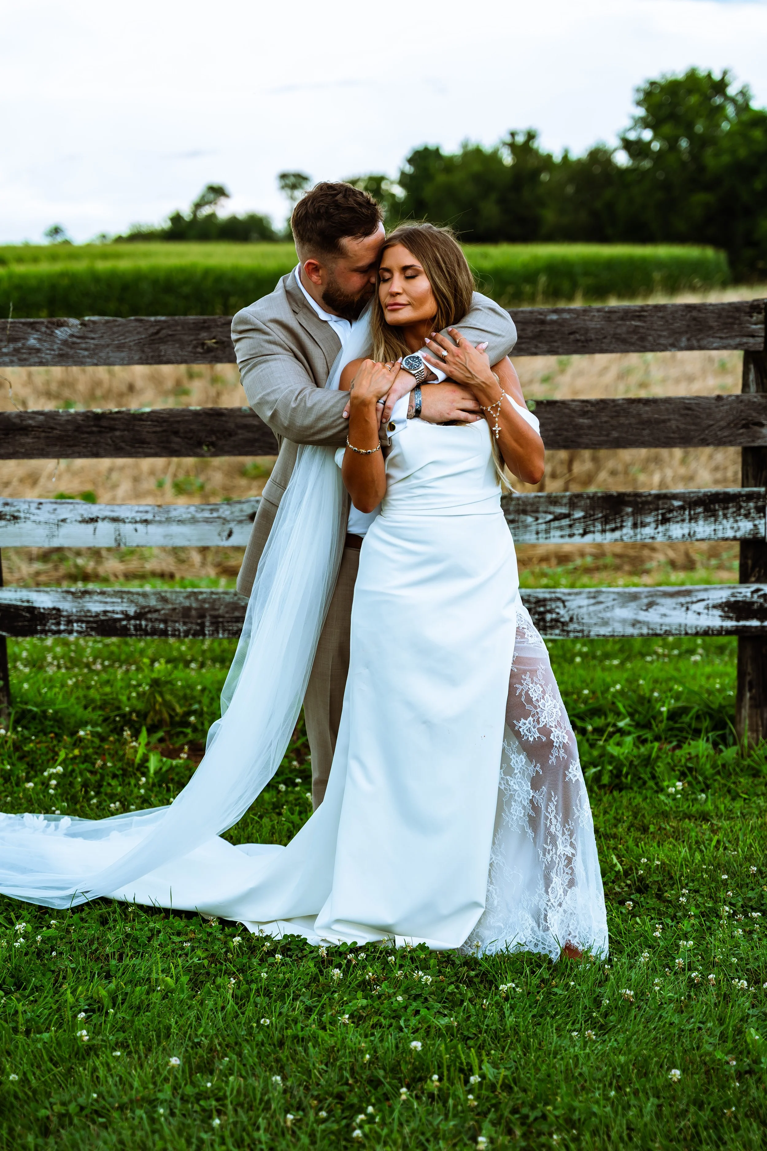 A bride and groom embrace outdoors near a wooden fence, with lush green fields and trees in the background. The bride is wearing a white wedding dress with lace details, and the groom is in a light gray suit. They appear to be posing for a romantic p