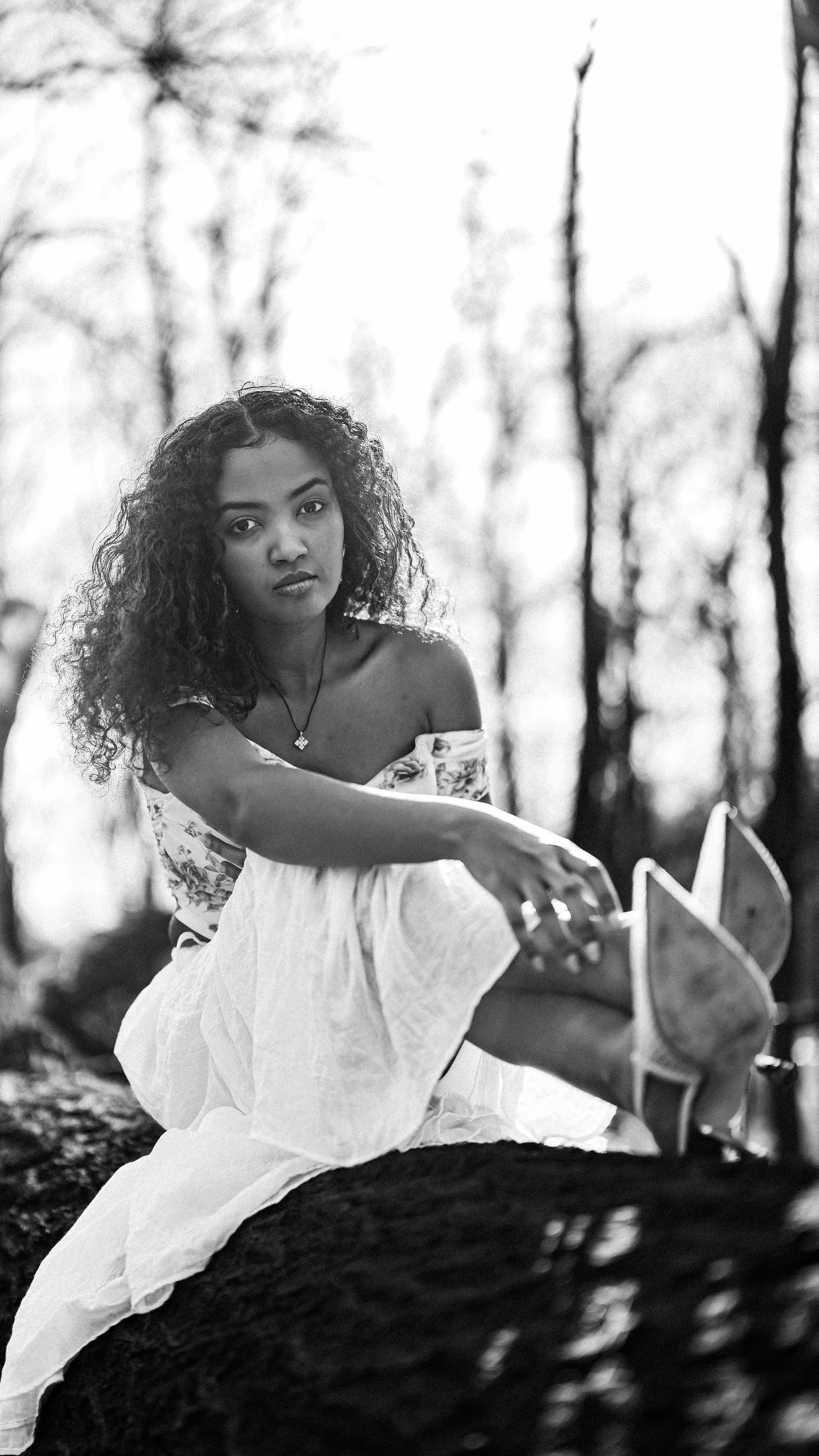 A woman with curly hair sitting on a fallen log outdoors, looking at the camera, with trees in the background.