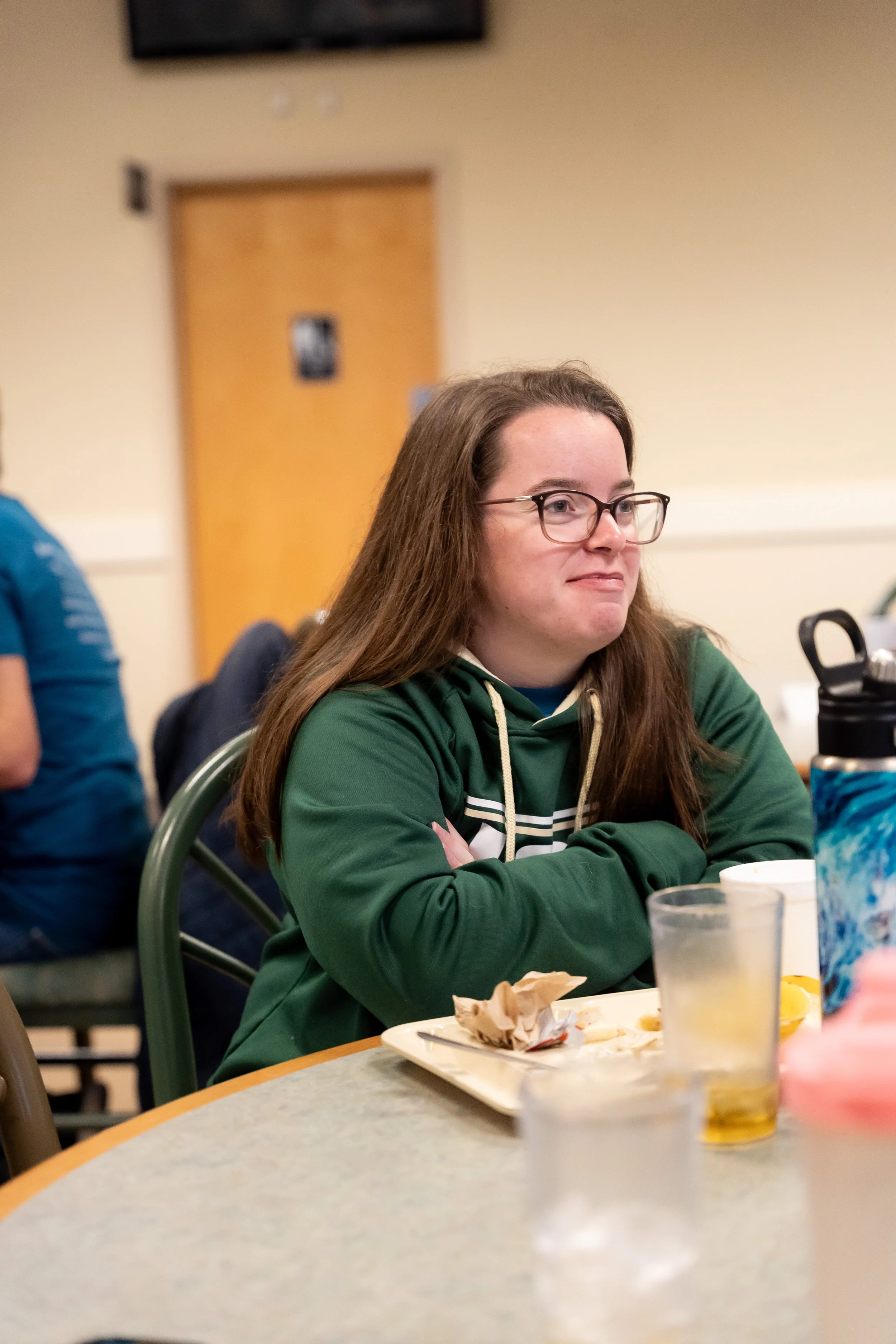 A woman with long brown hair, glasses, and a green hoodie sits at a table with a plate of leftover food, a glass of yellow drink, and a blue patterned water bottle, with a beige wall and door in the background.
