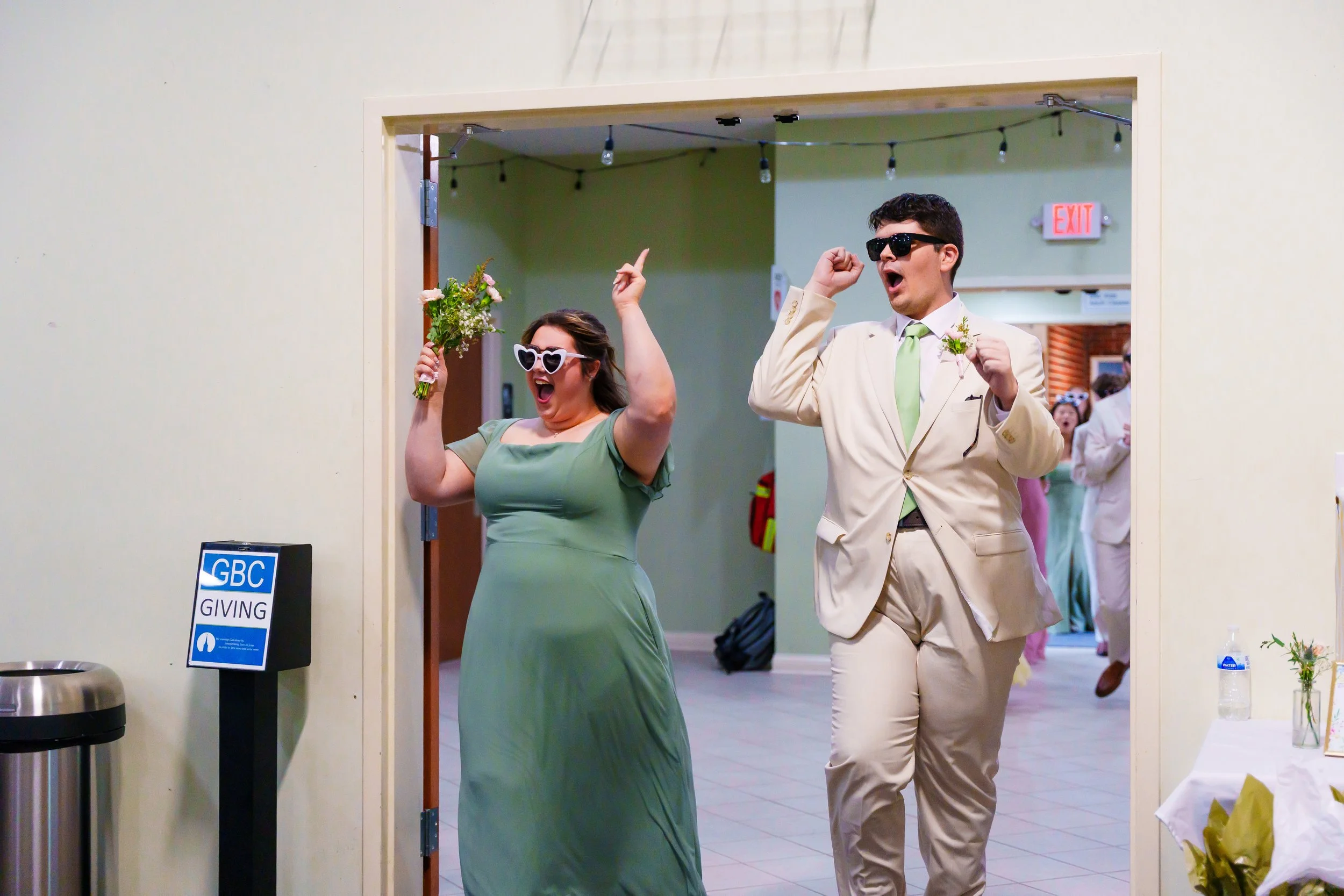 A joyous woman in a green dress holding flowers and wearing white sunglasses, dancing with a man in a beige suit with a green tie and dark sunglasses. They are celebrating at an indoor event, possibly a wedding reception.