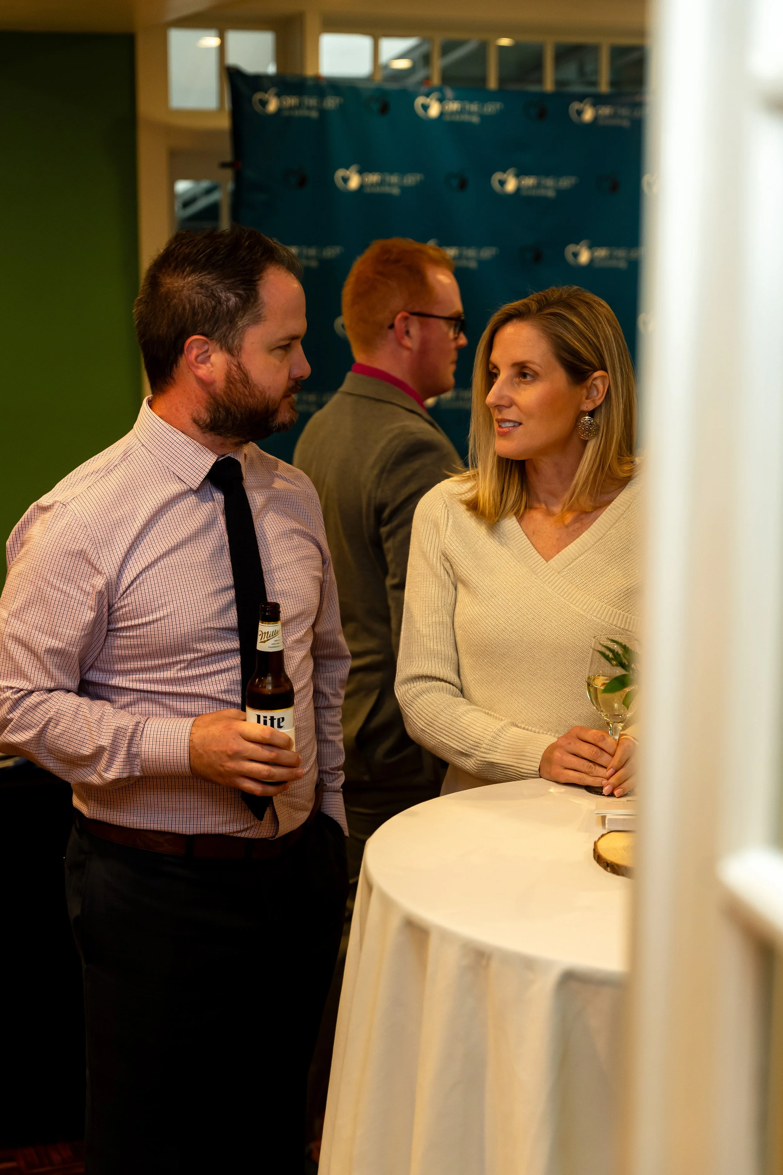 Two men and a woman engaged in conversation at a social event. The man on the left holds a beer bottle, the woman on the right holds a glass of wine, and a man in the background is blurred. They are standing near a white round table with a small tray