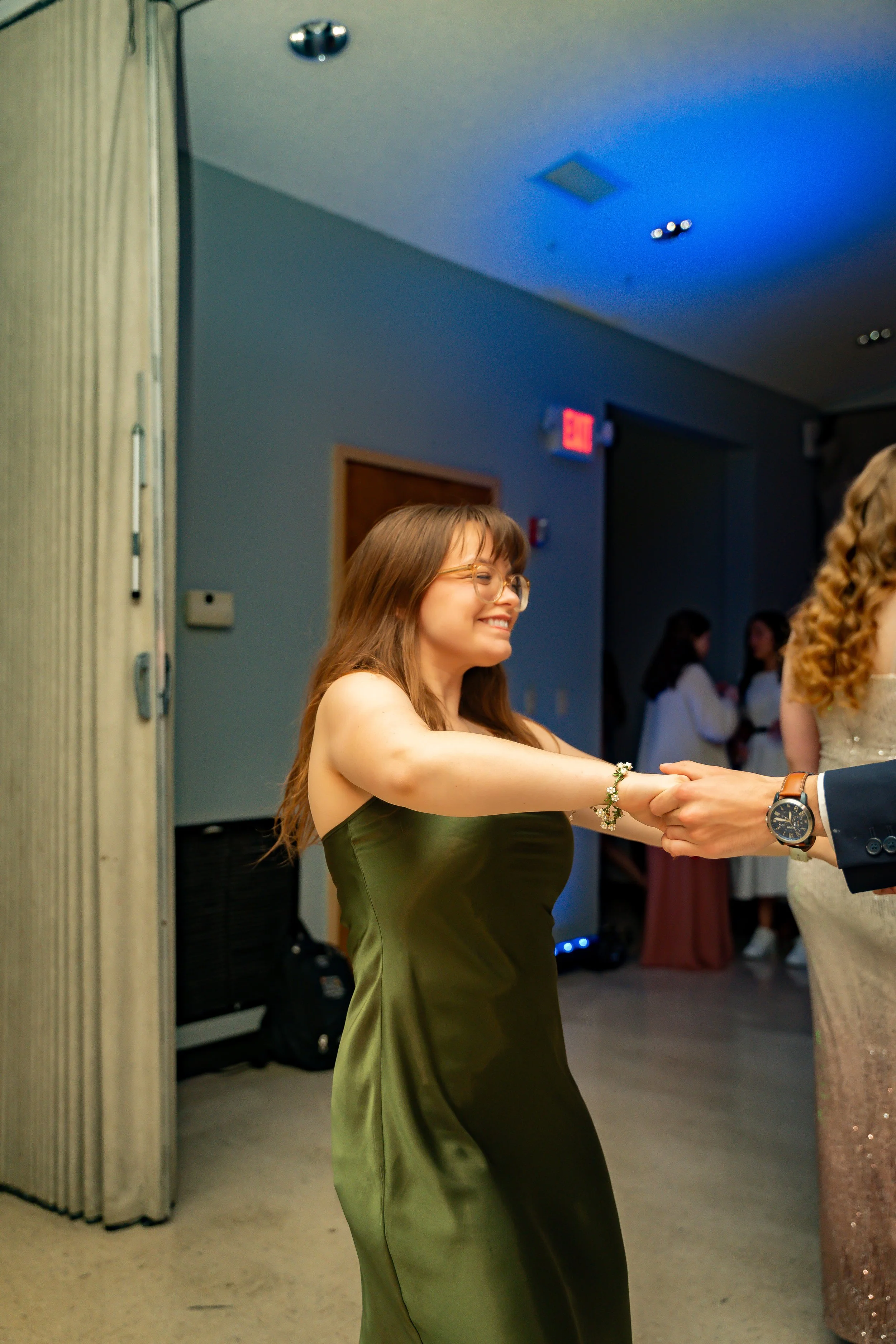 A woman in a green satin dress with glasses is smiling while holding hands with someone wearing a suit jacket. They are in a room with a dark wall, a door, and several other people in the background, some in formal attire.