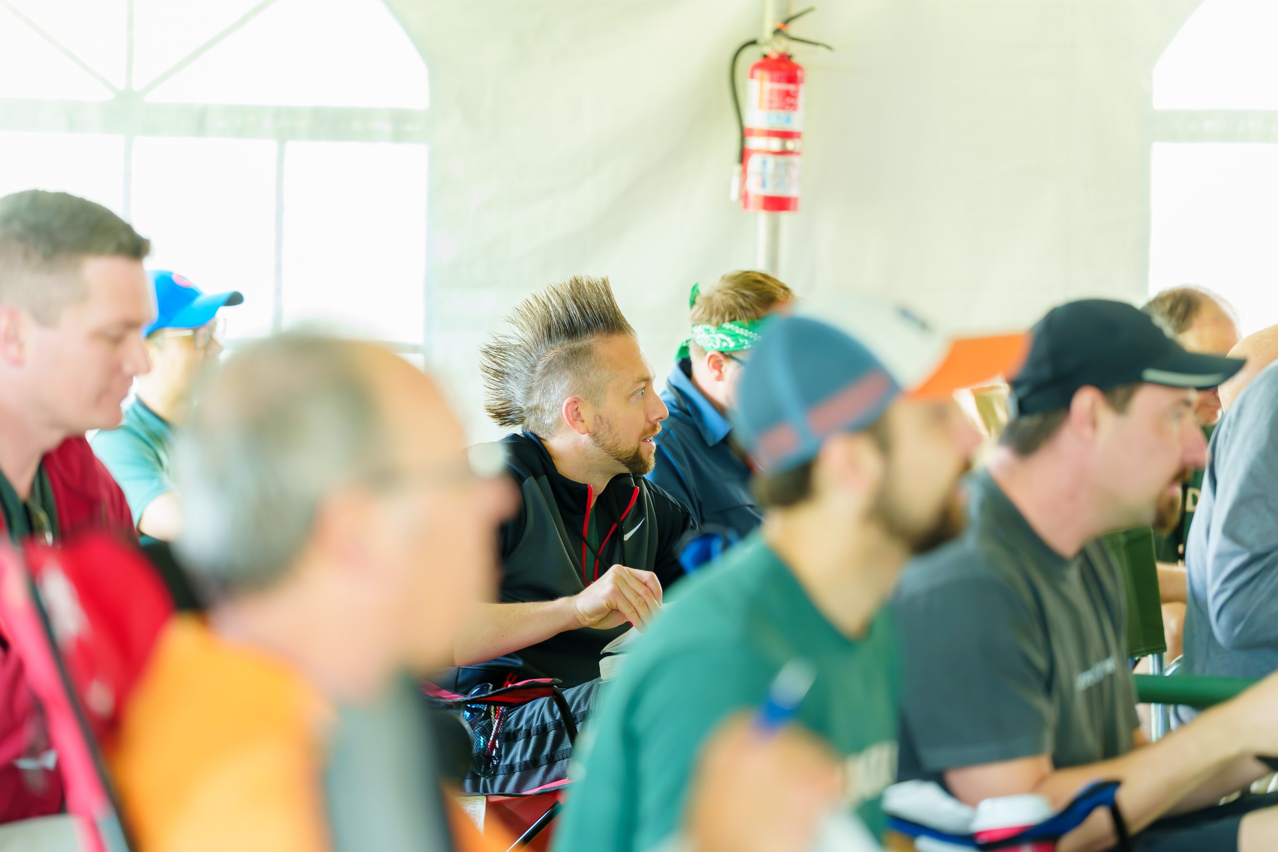 A group of people sitting indoors, attentively listening, with some wearing hats and bandanas, and a fire extinguisher mounted on the wall in the background.