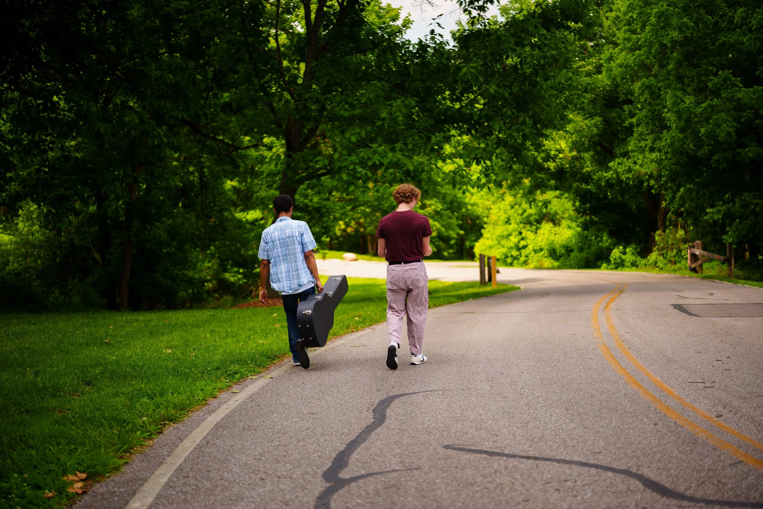 Two young men walking on a paved road in a lush green park, one carrying a guitar case, surrounded by tall leafy trees.