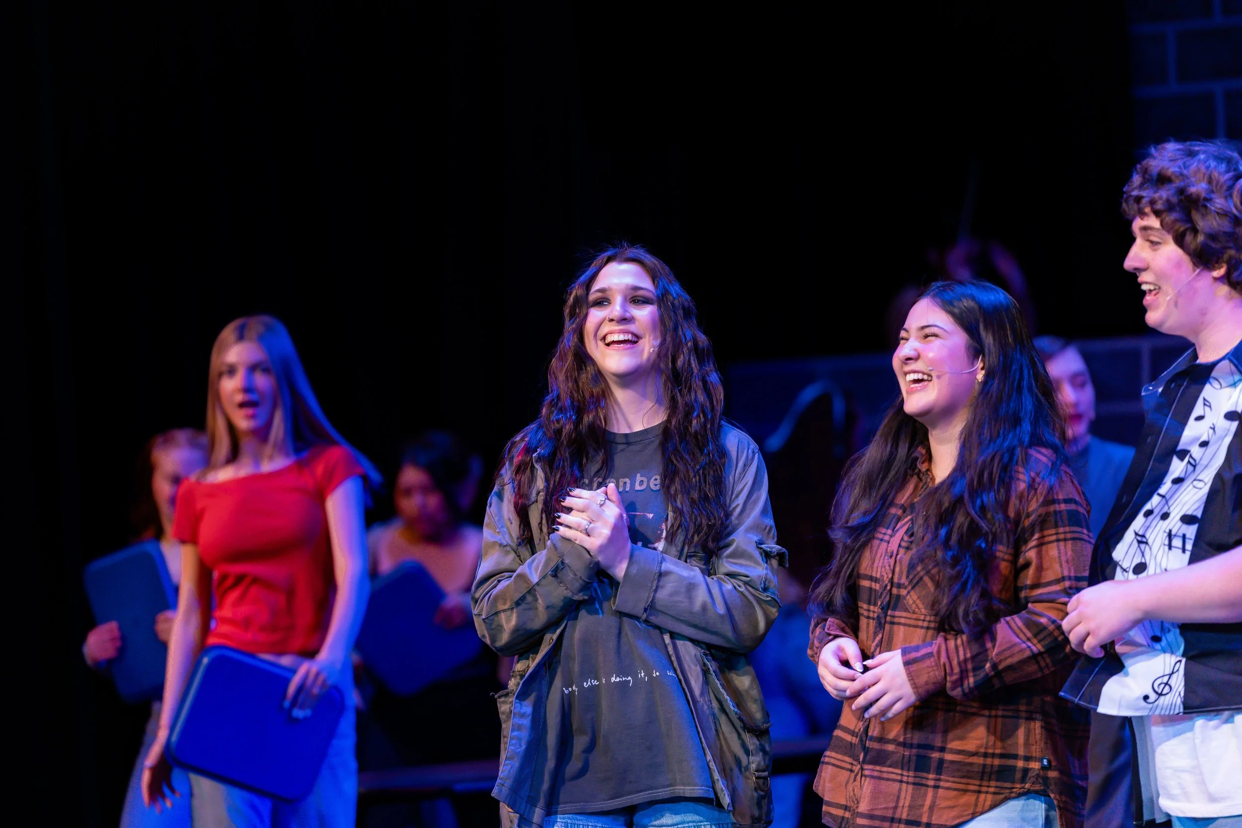 Group of young people smiling and engaging in a performance or rehearsal on stage, with some wearing microphones and music-themed clothing.