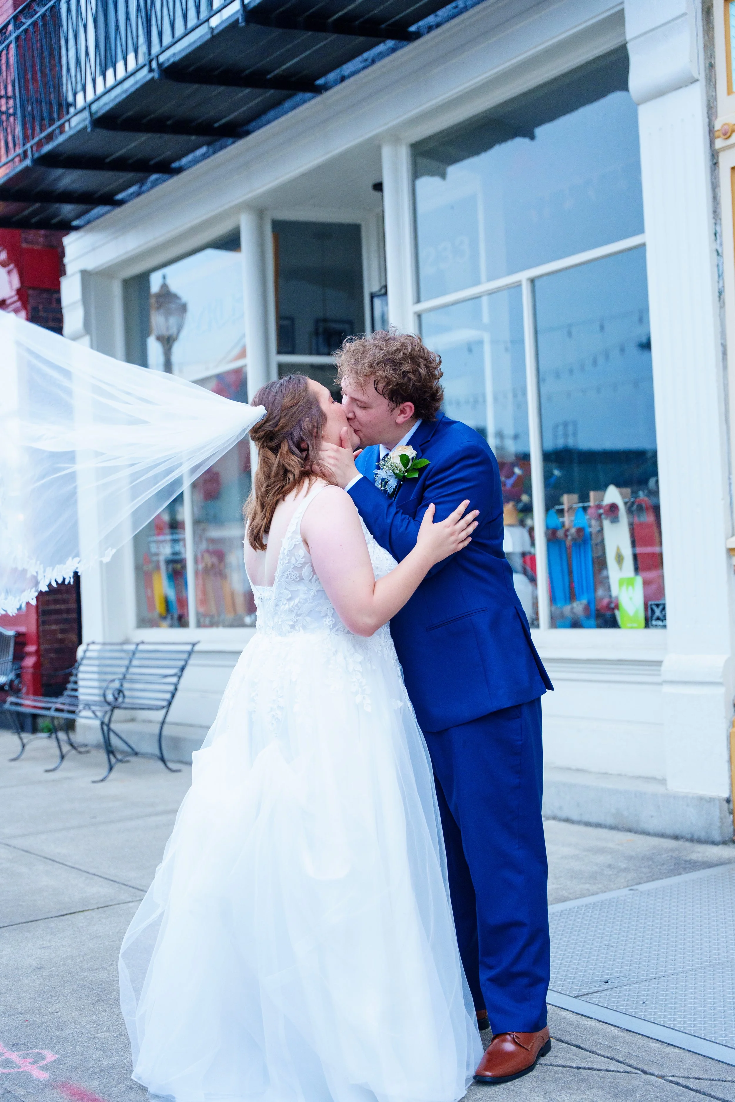 A bride and groom kissing outside a storefront, with the bride in a white wedding dress and the groom in a blue suit.