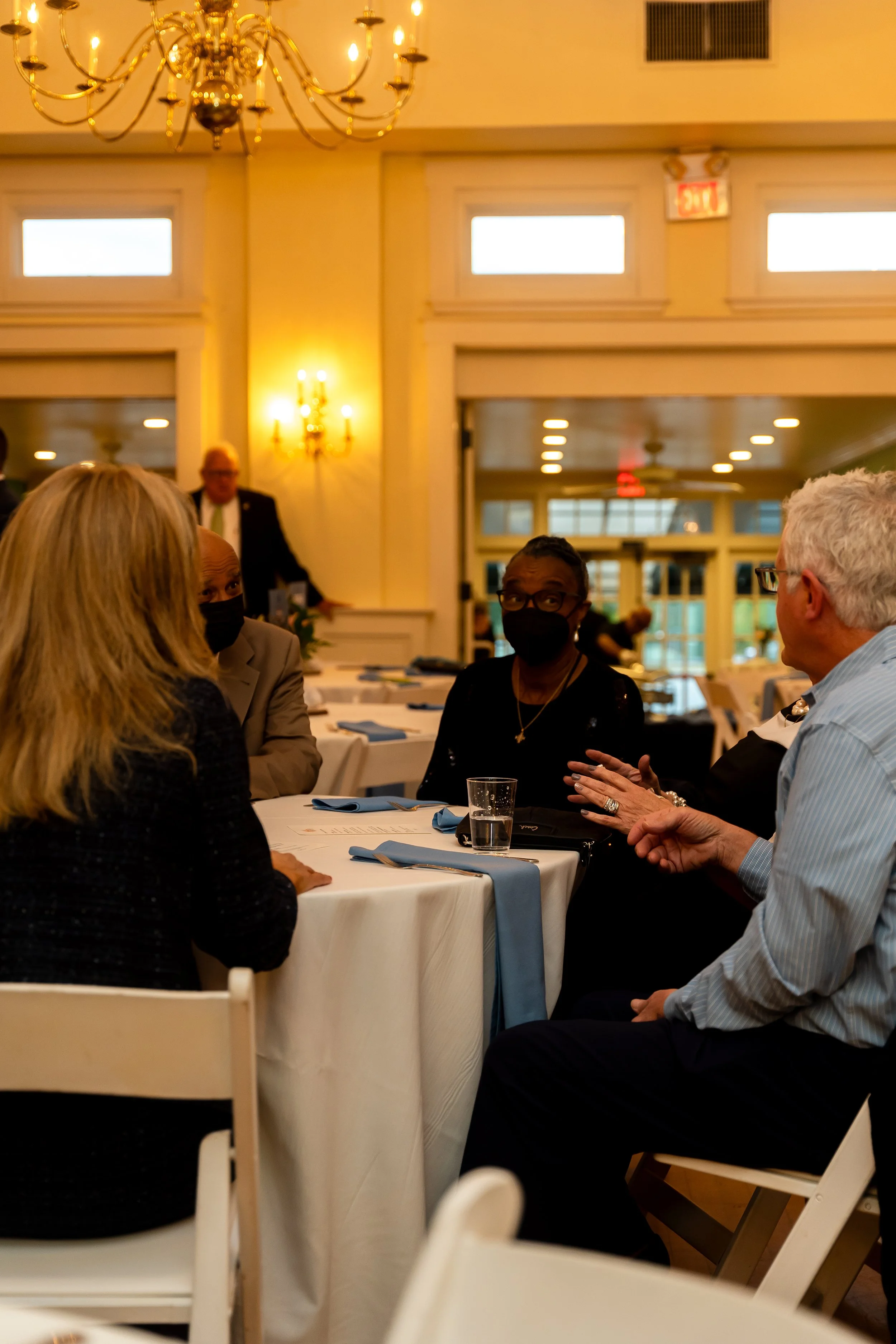 People sitting around a table in a formal event space, engaged in conversation, with a chandelier hanging from the ceiling, and large windows in the background.