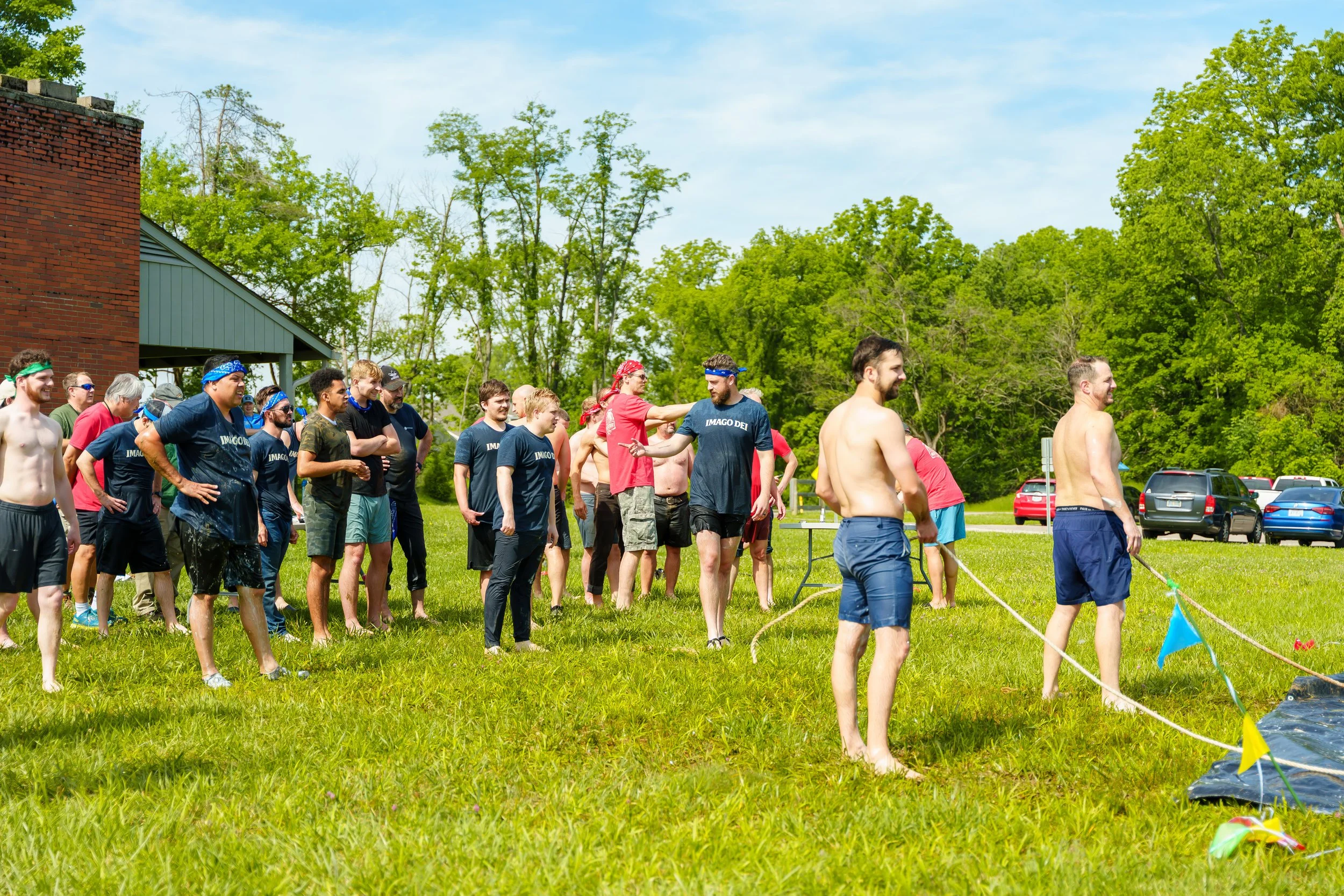 Group of people outdoors on a sunny day, participating in a water-based activity, with some wearing headbands and casual clothing, standing on a grassy area near a building and trees.