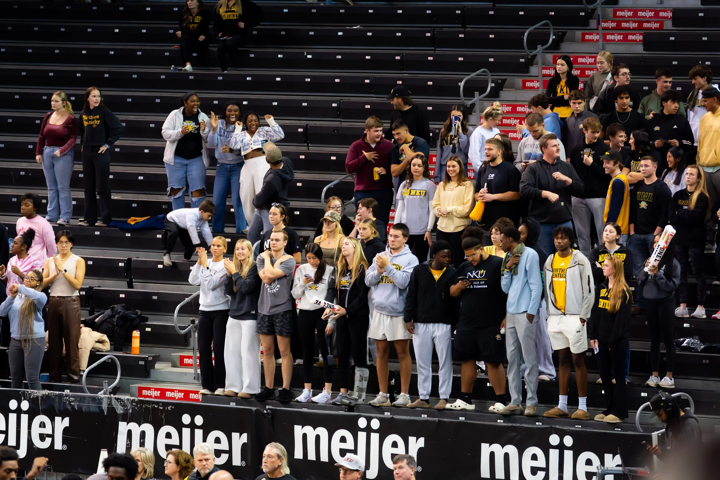 Group of people, mostly young adults, standing and sitting in stadium bleachers, some wearing athletic clothing and school apparel, with a few holding signs and engaging in conversations.