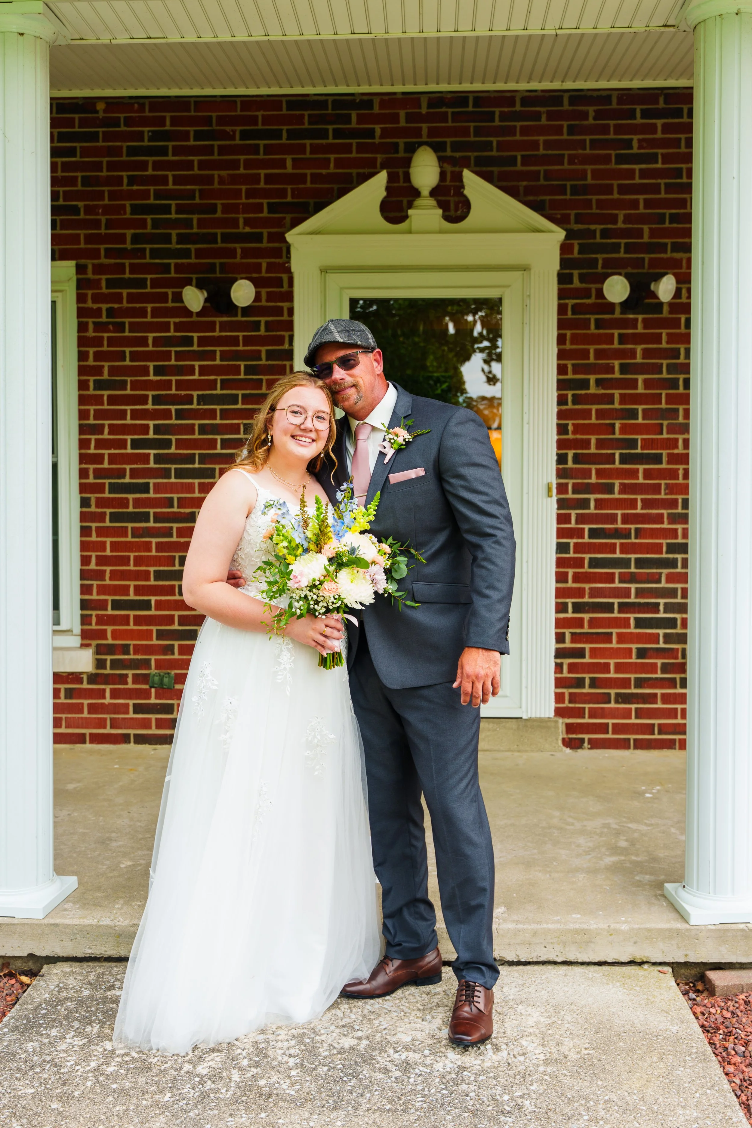A bride and groom stand close together outside a house with red brick walls and white columns. The bride is wearing a white wedding dress and glasses, holding a bouquet of colorful flowers. The groom is wearing a dark suit, pink tie, and sunglasses. 
