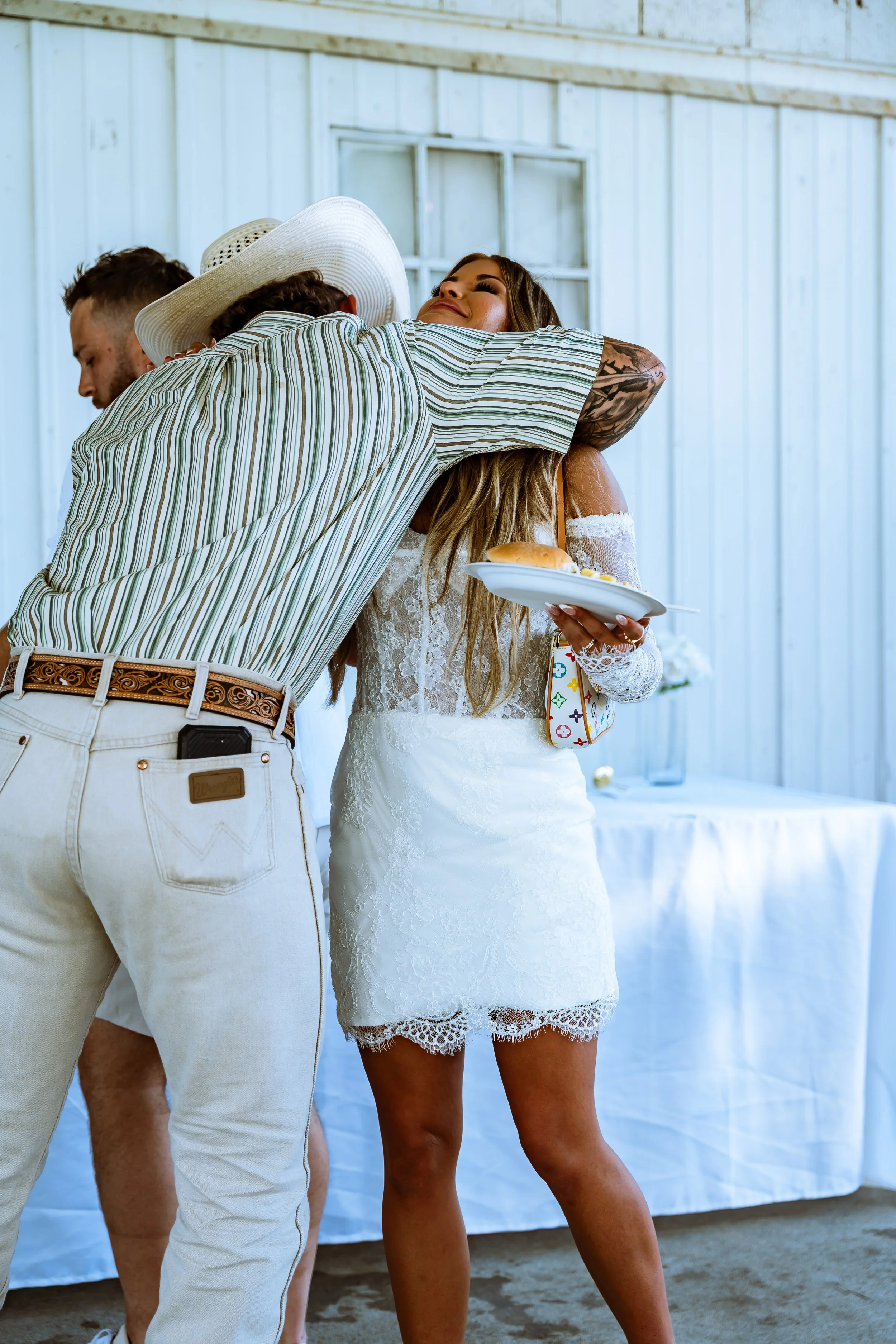 A man in a striped shirt and white pants hugging a woman in a white lace dress with a tattoo on her arm, while holding a plate of food, at an outdoor event with a white wooden wall in the background.