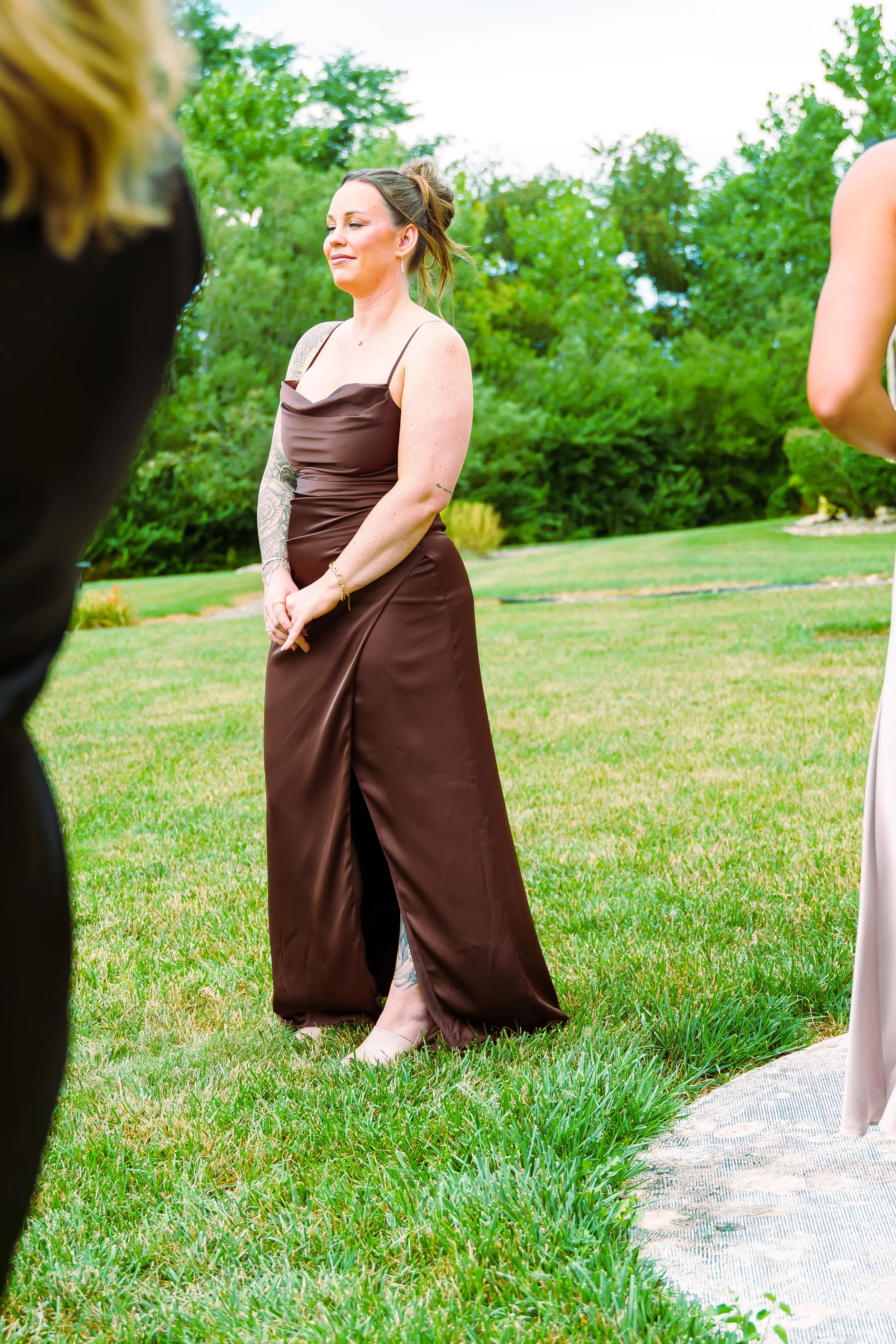 A woman in a brown satin dress standing outdoors on a lawn with trees in the background, smiling with her eyes closed.