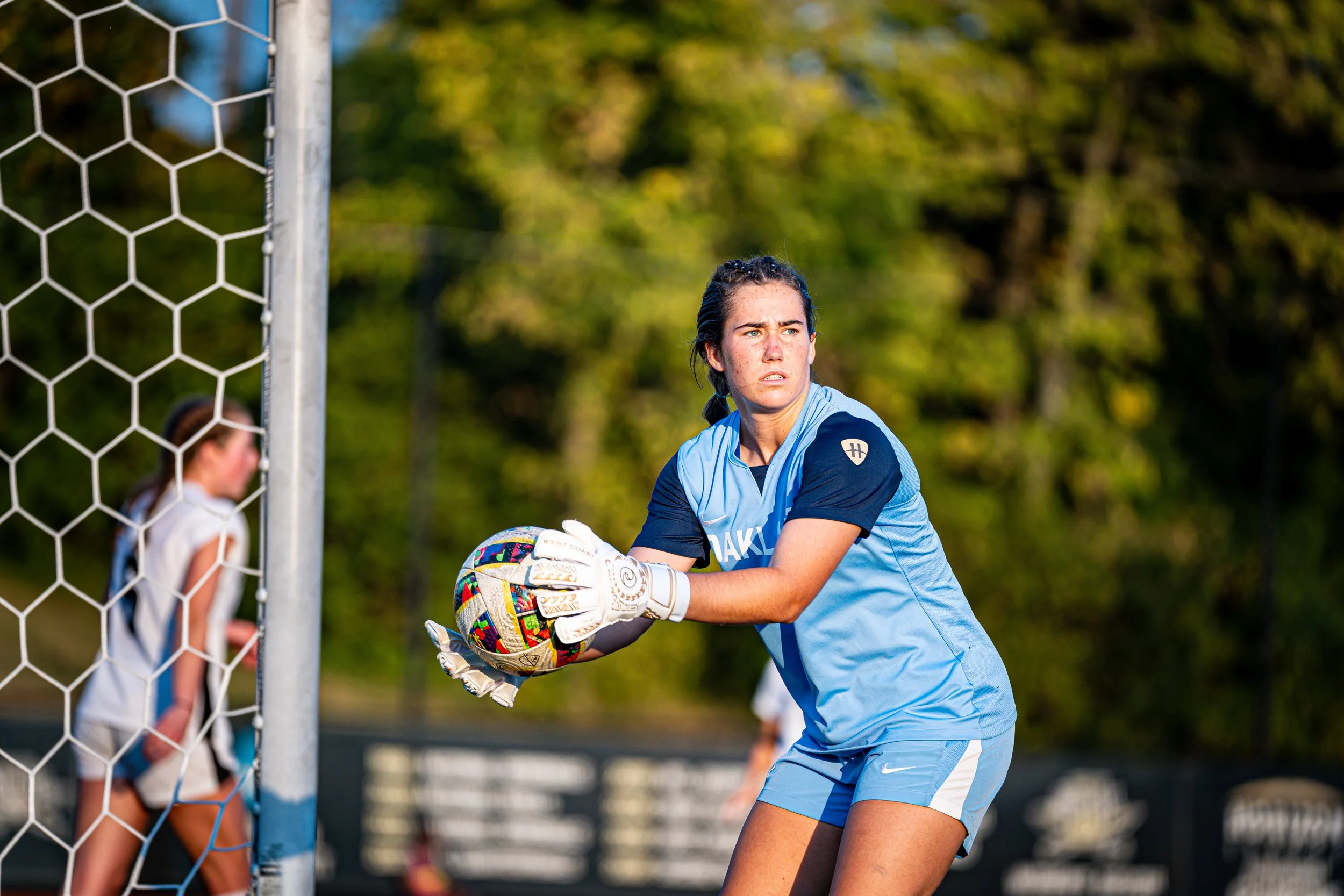 A female soccer goalkeeper in a light blue uniform with black sleeves, wearing goalie gloves, holding a colorful soccer ball during a match on a grassy field with a net, other players, and trees in the background.