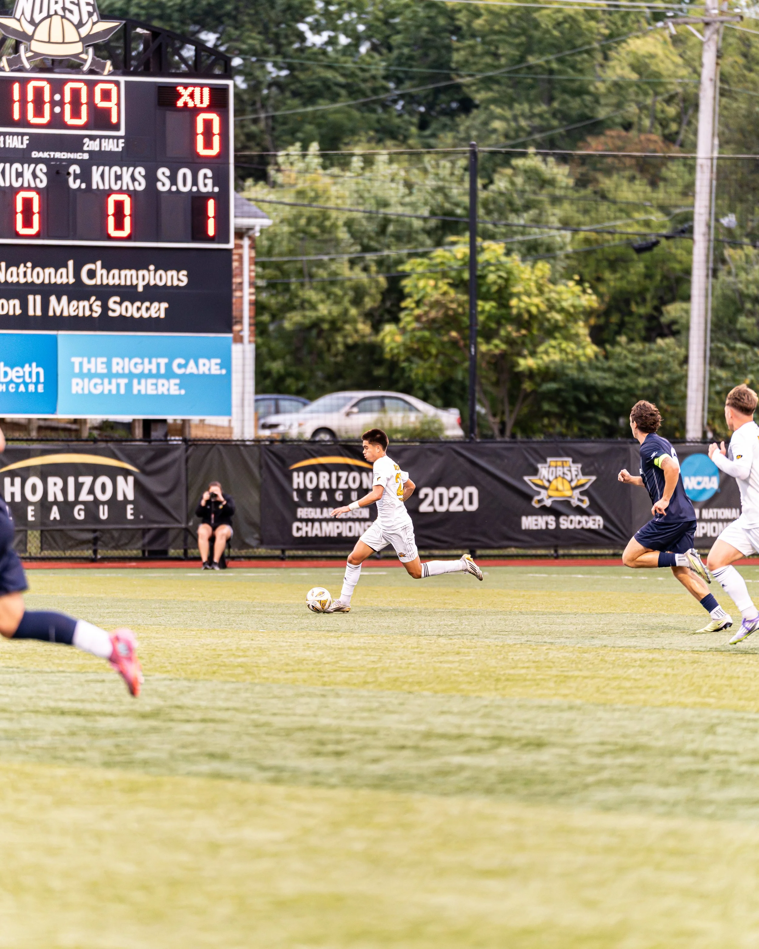 A youth soccer game in progress on a field with players running and a player in white controlling the ball. The scoreboard shows 10 minutes and 9 seconds remaining in the first half, with the score tied at 0-0. There are trees and parked cars in the 