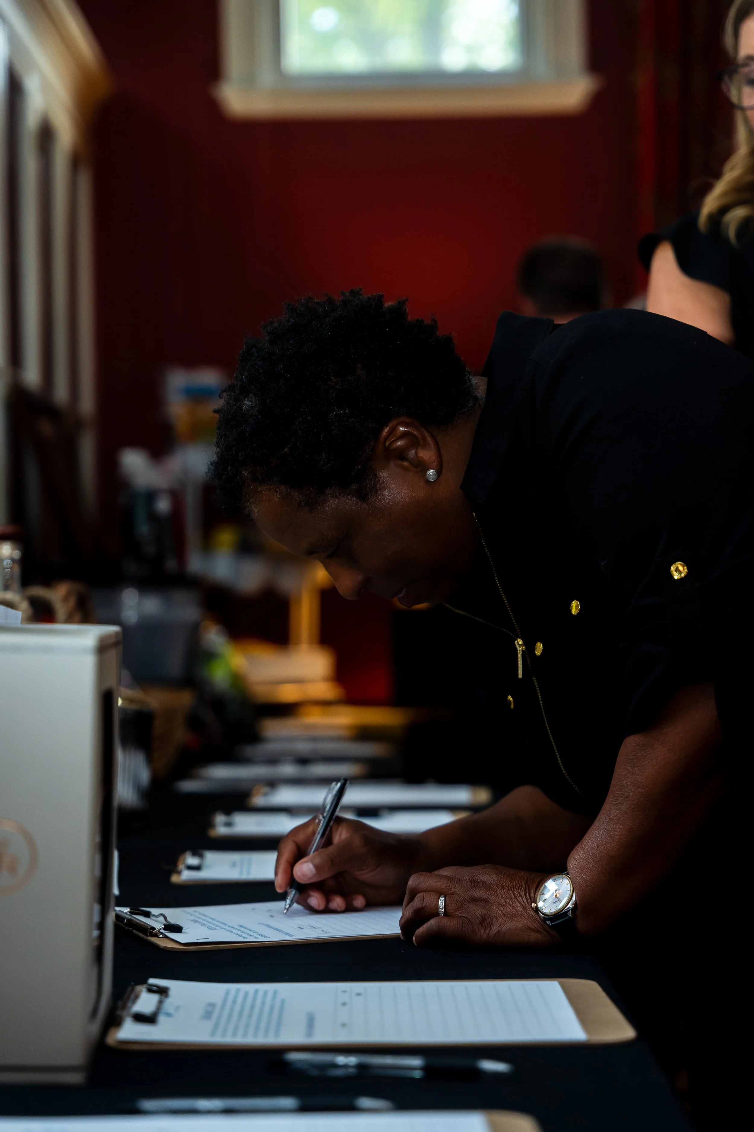 A person with dark curly hair and a wristwatch, signing a document on a clipboard at a table, with other documents and office supplies nearby, in a room with red walls and a window.