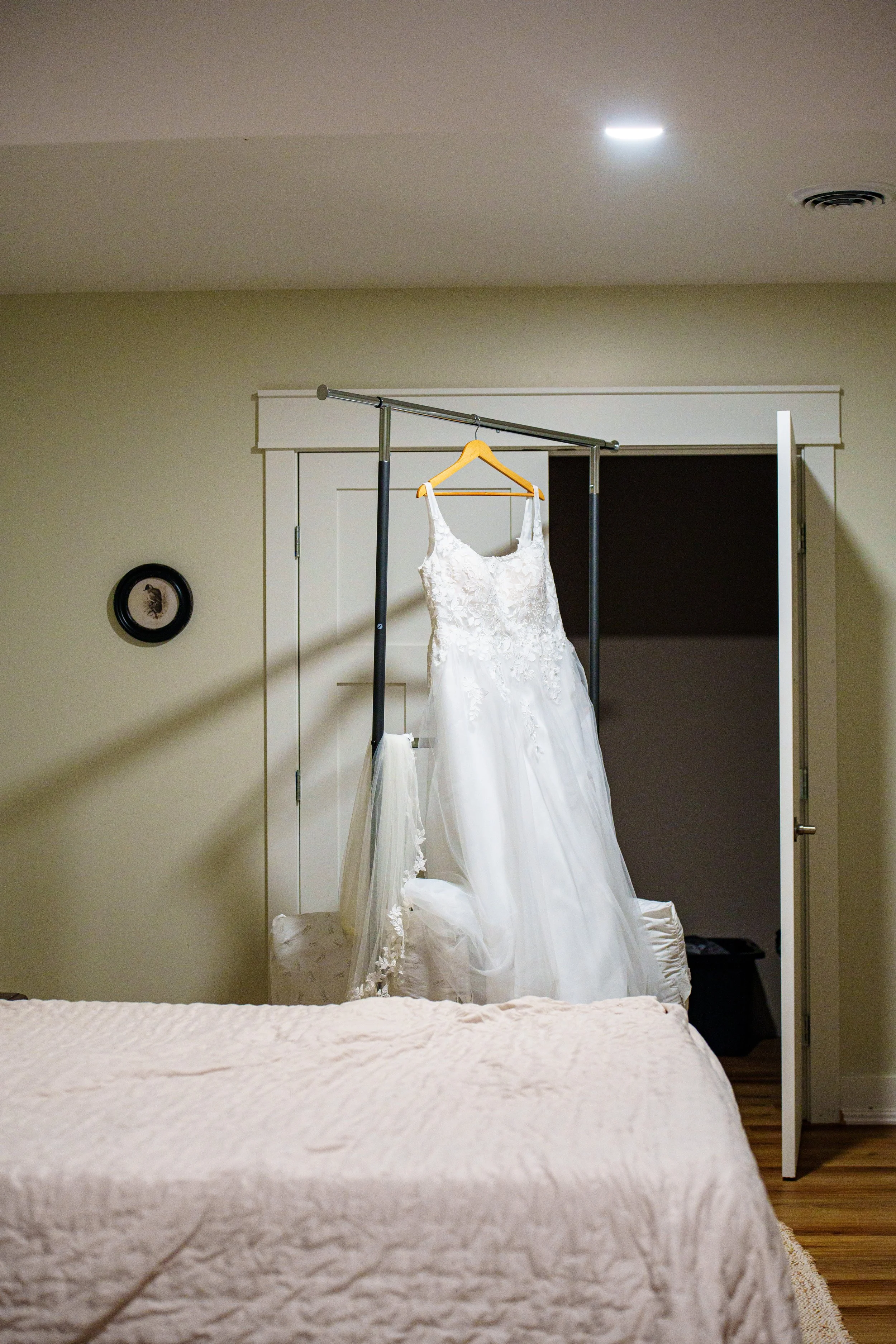 Wedding dress hanging on a clothing rack in a bedroom with a closed door and wall decor.