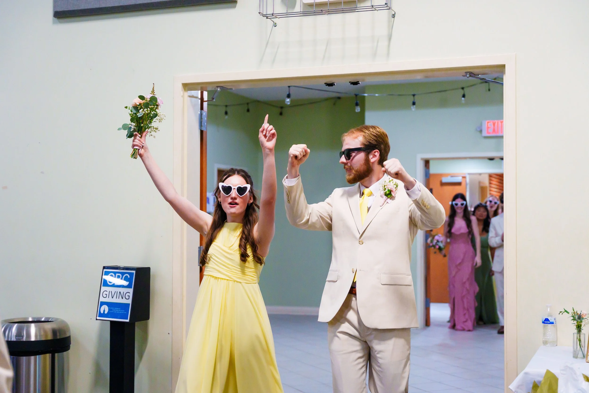 A woman in a yellow dress wearing heart-shaped sunglasses holding a bouquet of flowers with her arm raised, and a man in a cream suit with a boutonniere raising his arm, both celebrating inside a building with other people in the background.