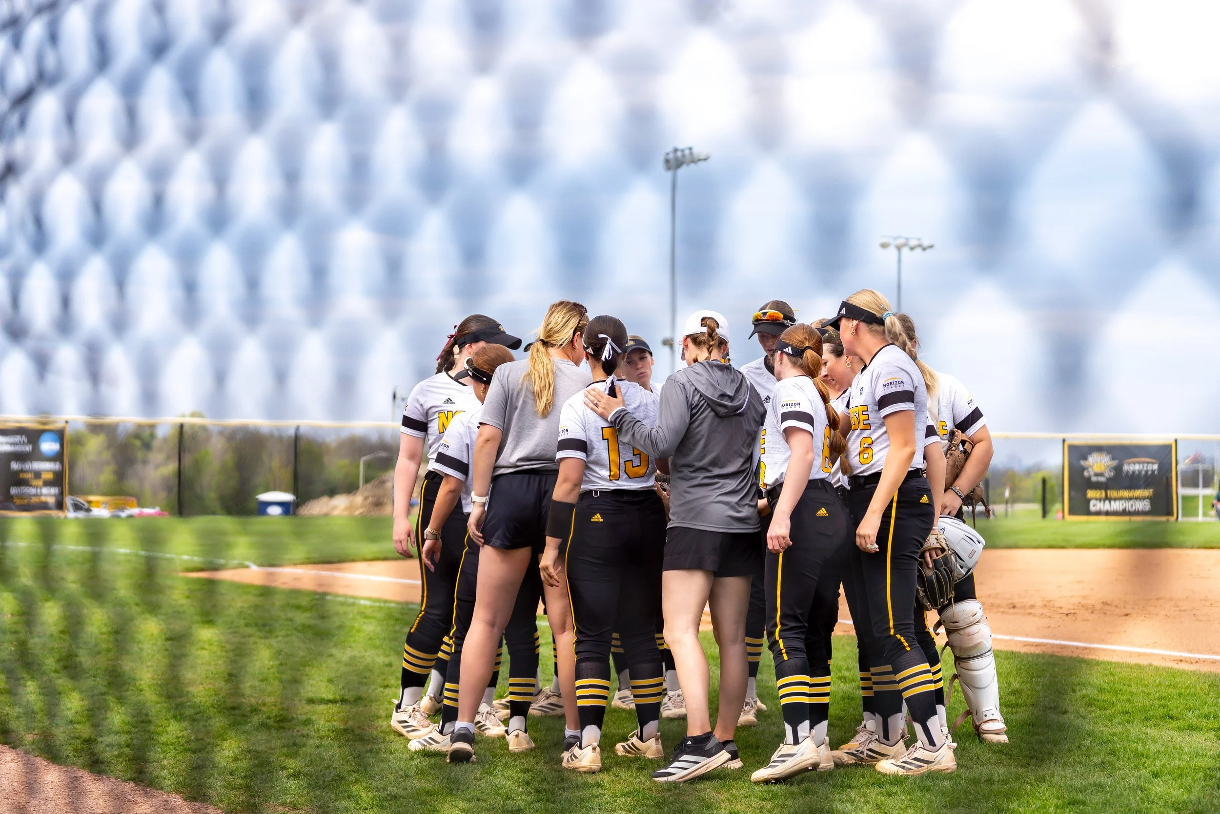 A softball team in black, white, and yellow uniforms huddles together on the field during a game, with a large, modern stadium in the background.