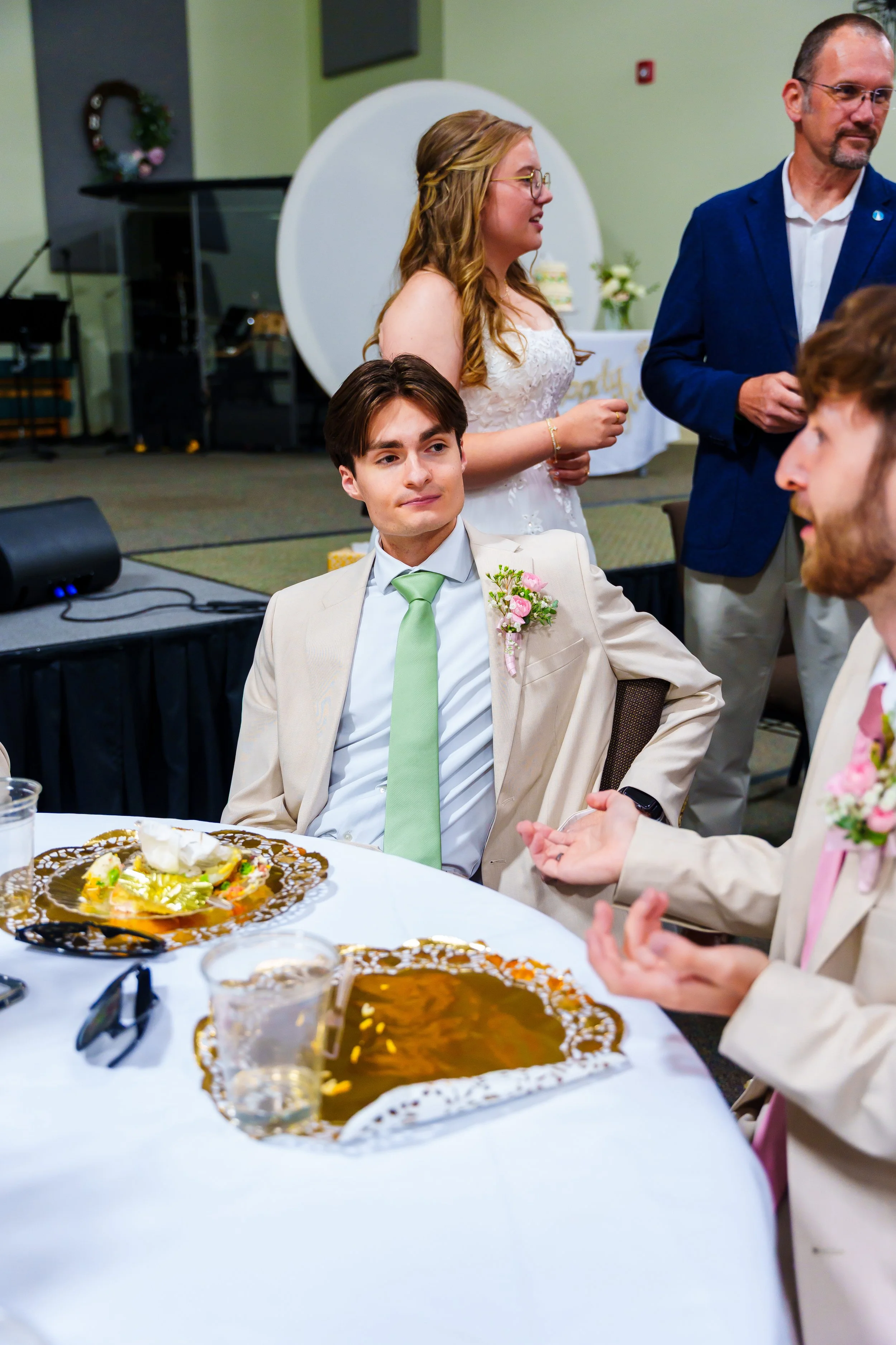 People sitting and standing around a table at a celebration or party, with food and drinks on the table, and a decorated background.
