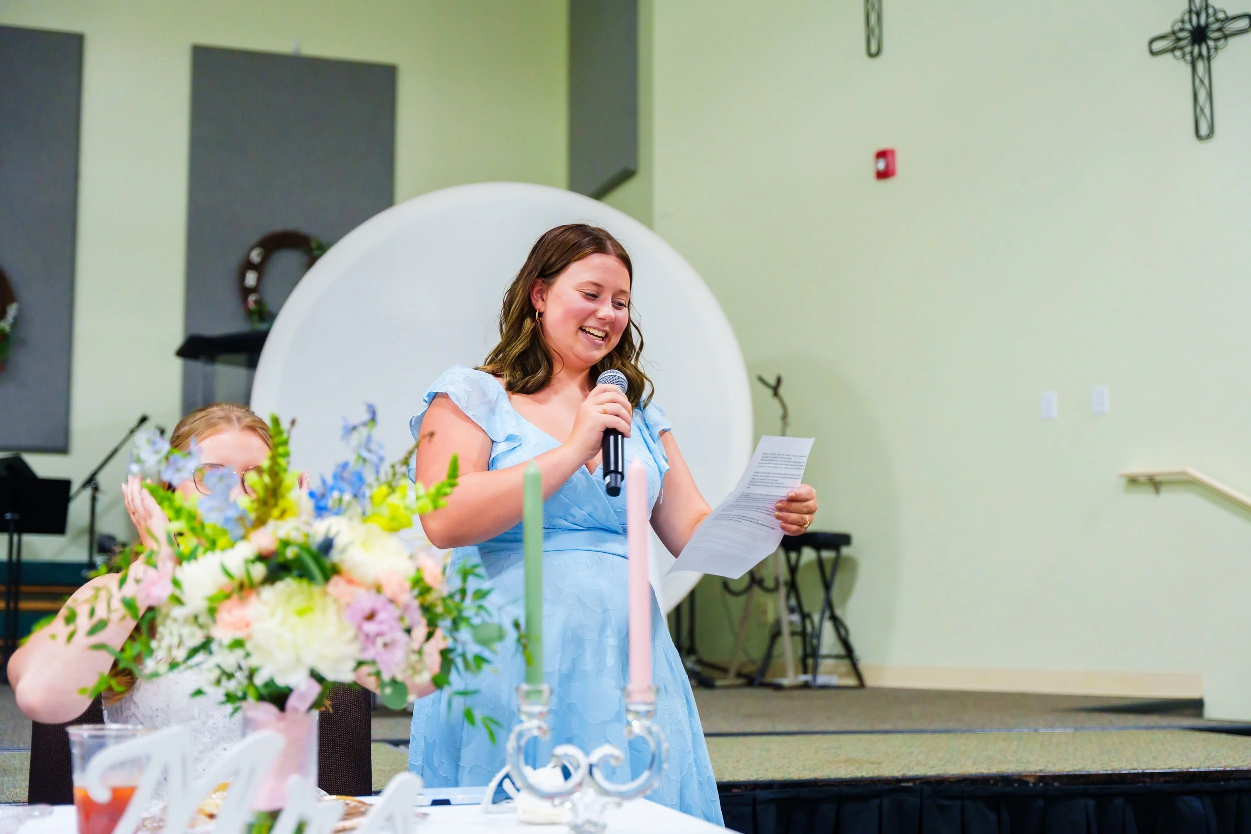 A young woman in a light blue dress speaking into a microphone during a celebration or ceremony, with floral arrangements and candles on a table in front of her.
