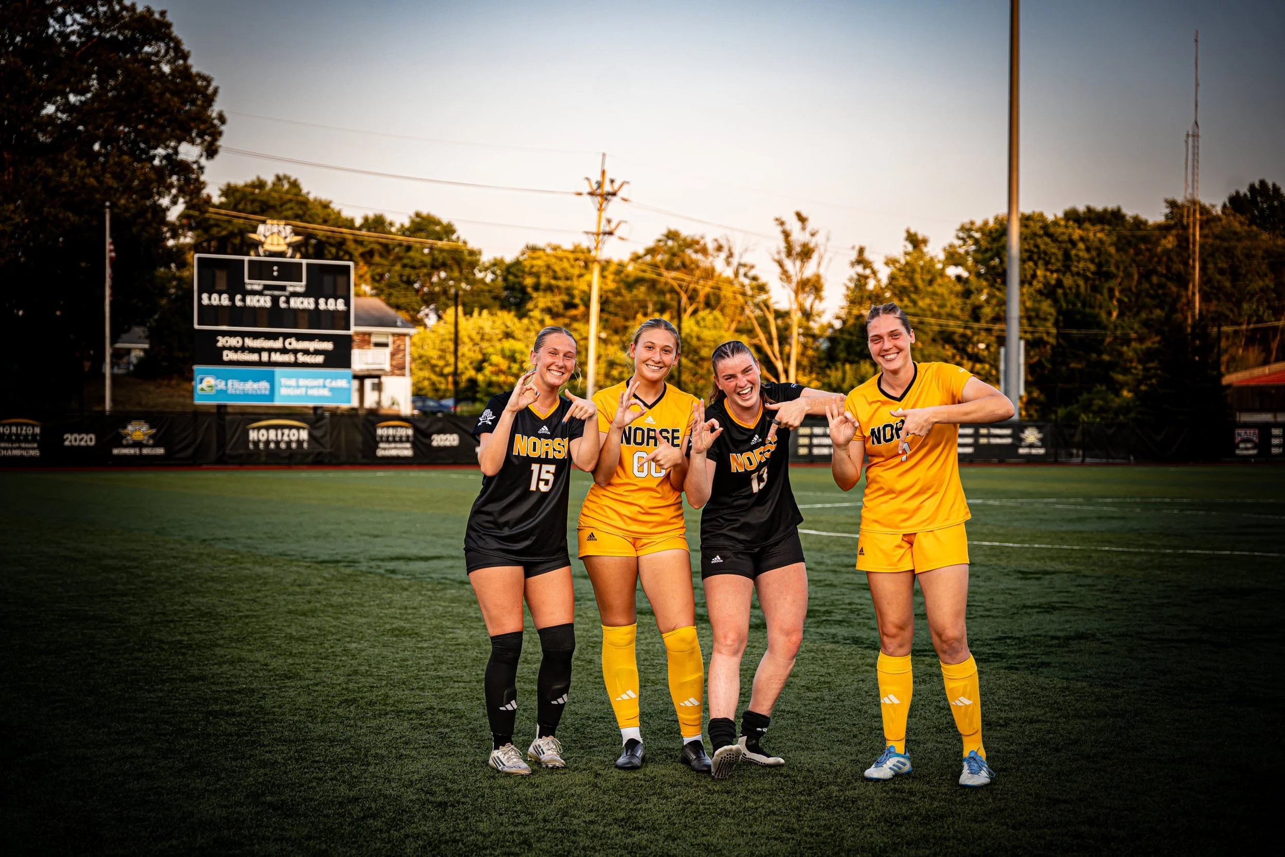 Four female soccer players in black and yellow uniforms standing on a soccer field, smiling and making celebratory gestures.