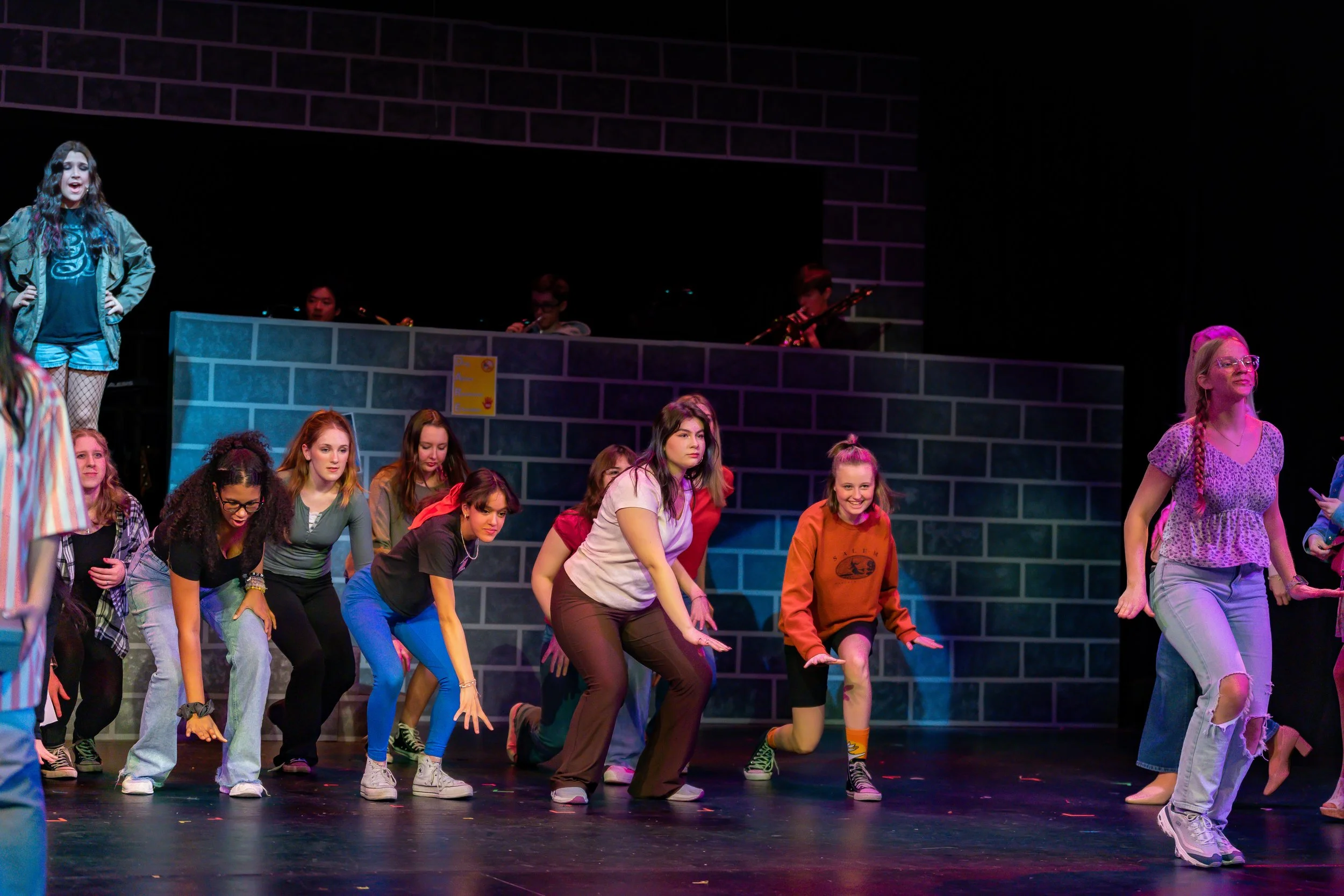 A group of young women perform a dance on a stage, with a dark backdrop and a band playing in the background.