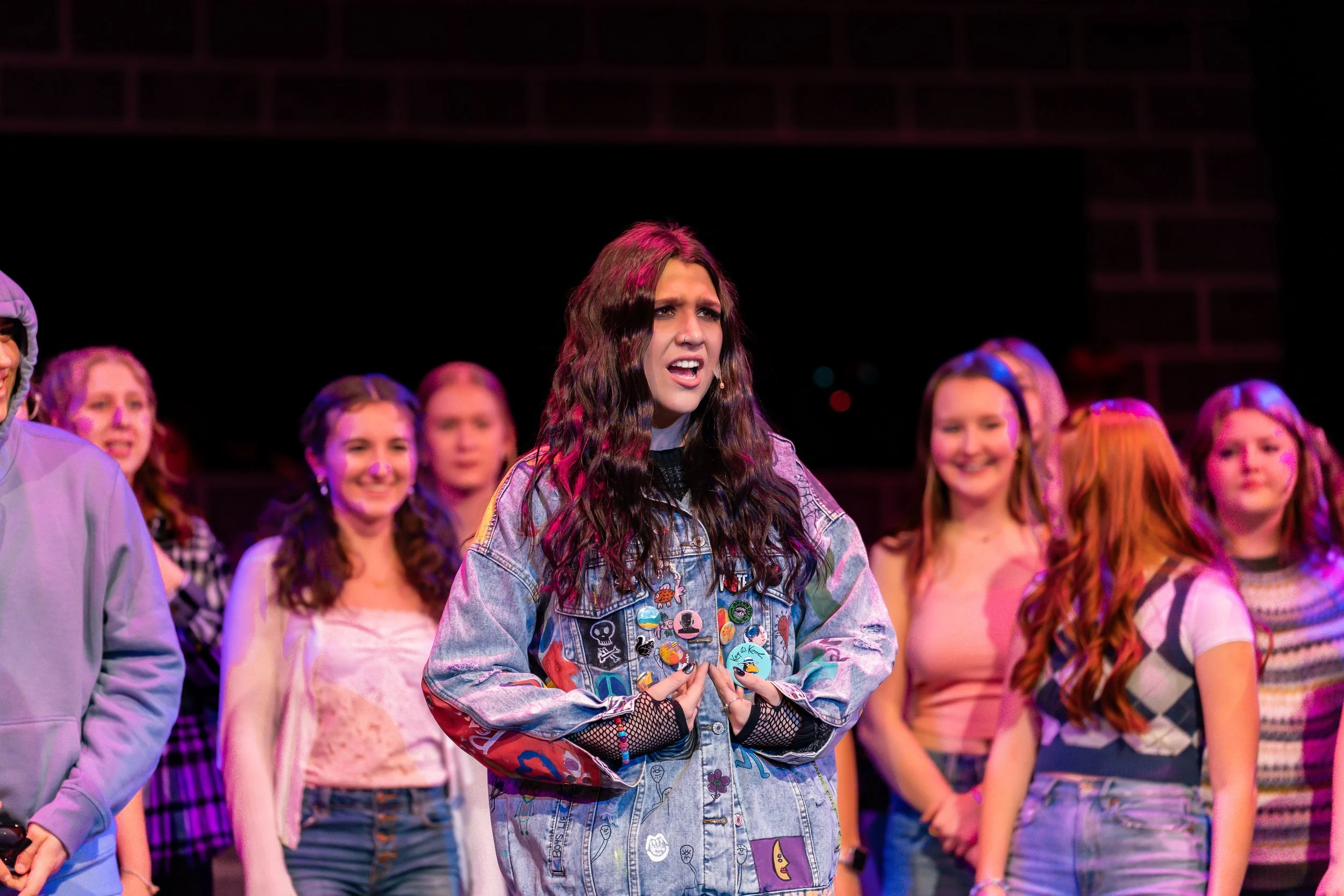 A group of young women standing together on a stage, with the woman in the center appearing to be speaking or singing. She has long, wavy dark hair and is wearing a denim jacket with various patches and buttons. The women surrounding her are smiling 