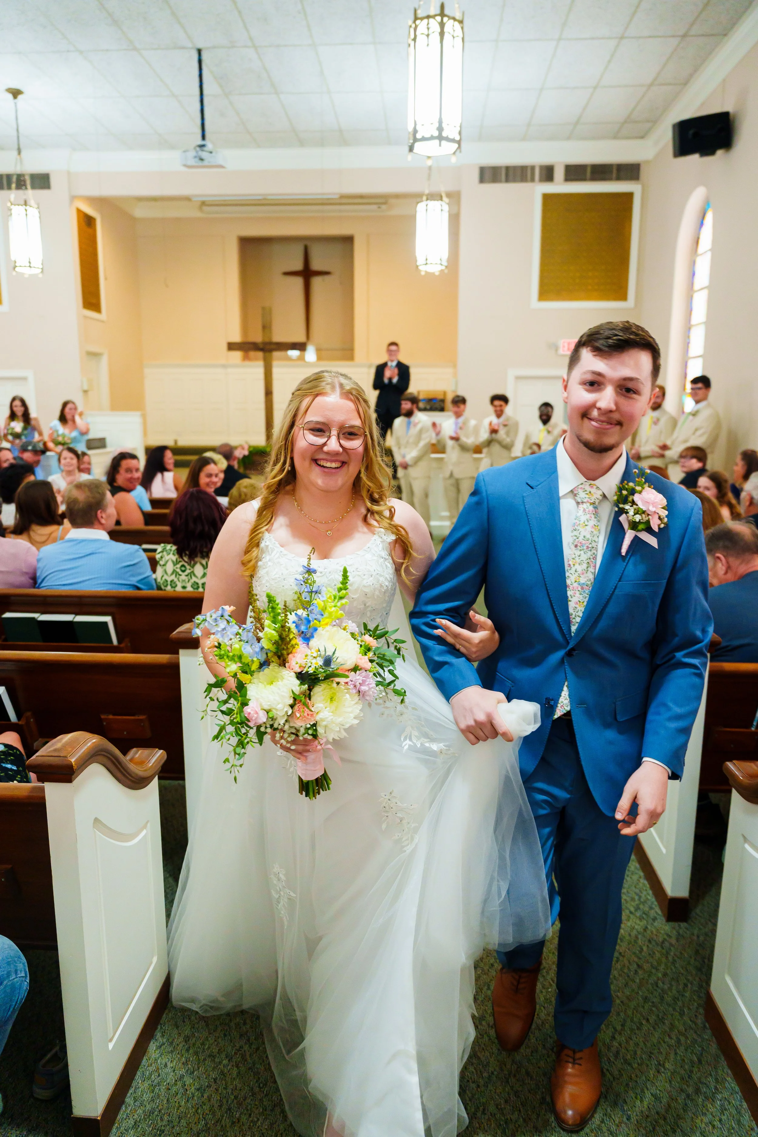 A bride and groom walk down the aisle arm in arm in a church during their wedding ceremony. The bride holds a large bouquet of colorful flowers and wears a white wedding dress. The groom wears a blue suit with a floral tie and brown shoes. Guests are seated and smiling in the background, with some standing at the front.