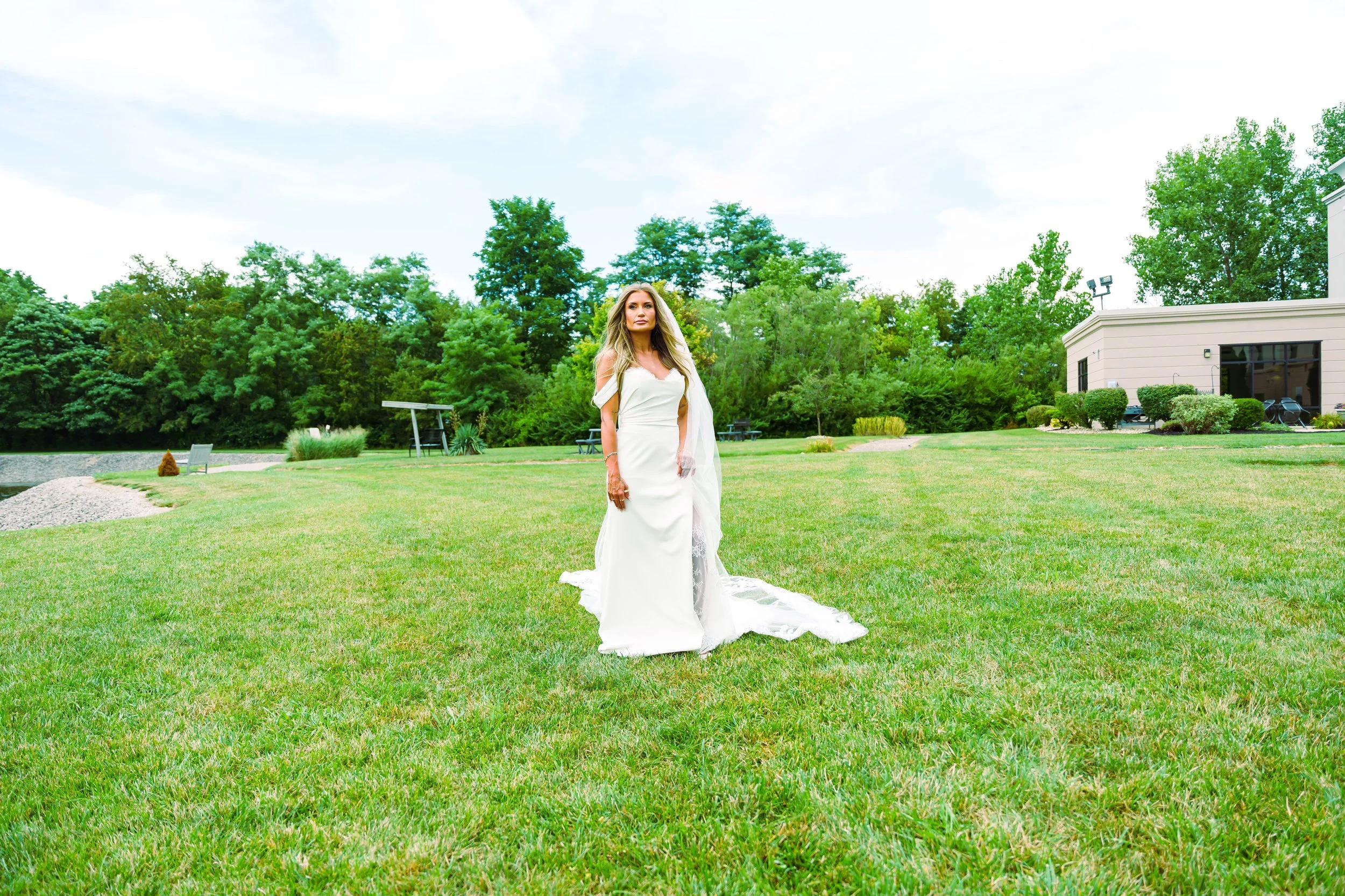 A woman in a white wedding dress standing on a well-kept lawn with trees and a building in the background.