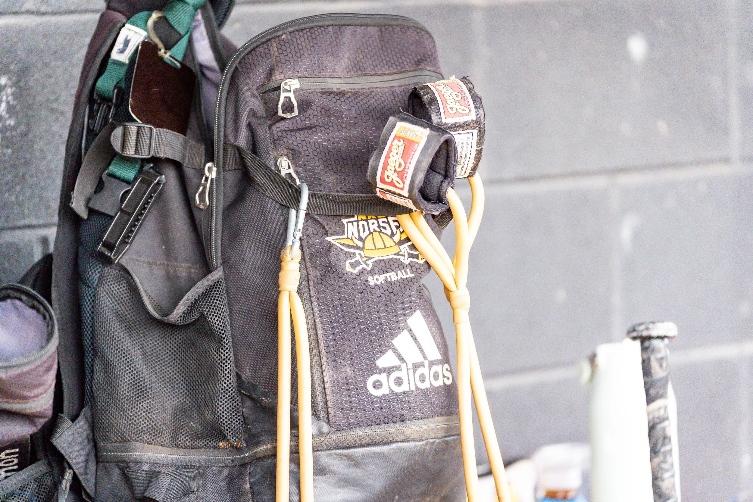 Close-up of a black Adidas sports bag with a softball team's logo, a pair of yellow crutches hanging on it, and a portion of a backpack with mesh pockets and zippers, against a gray background.
