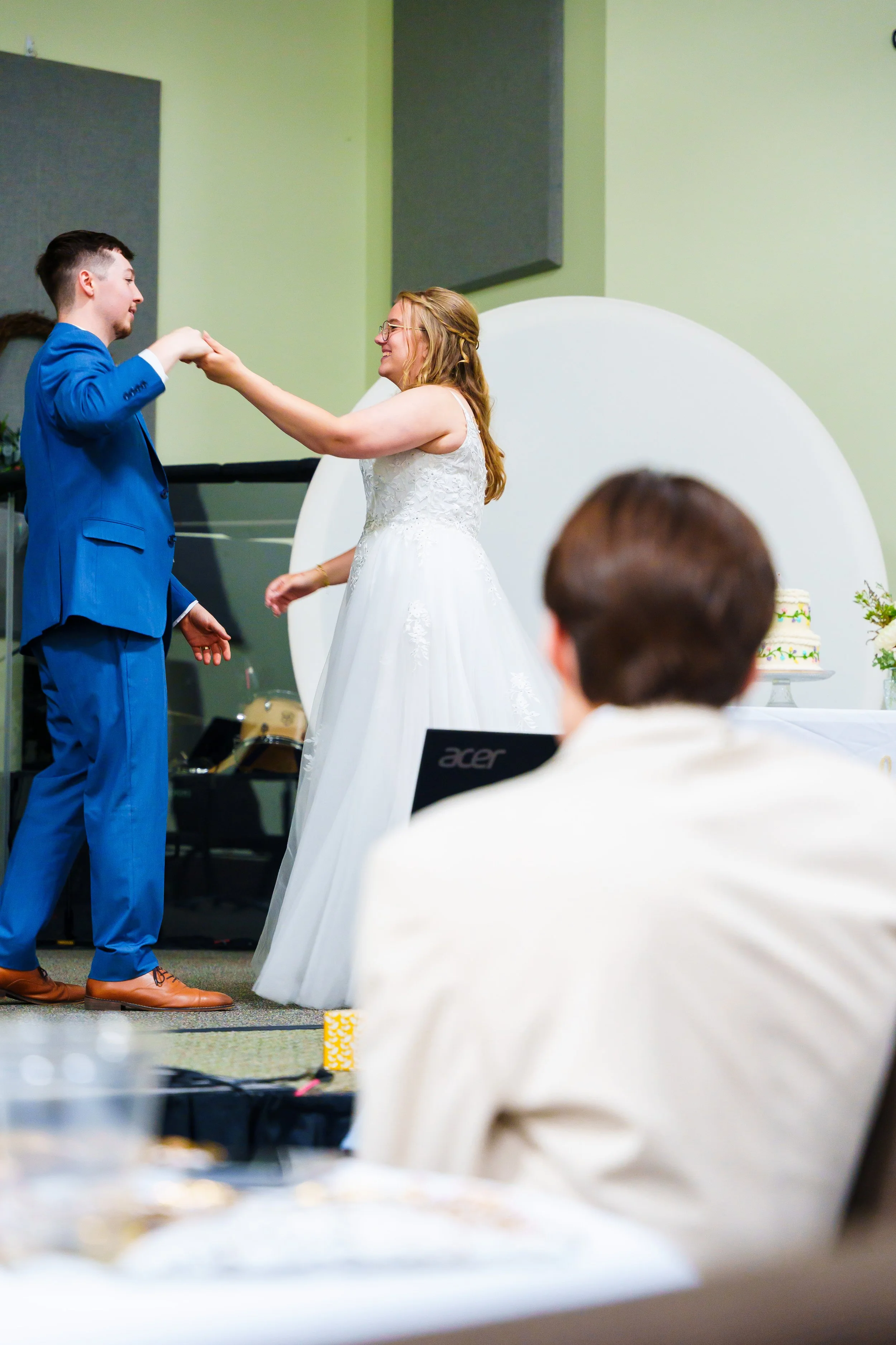 A bride and groom dancing at their wedding reception, with the bride wearing a white dress and the groom in a blue suit. They are smiling and holding hands, with a cake visible on a table nearby. The scene is indoors with green and gray walls and an 