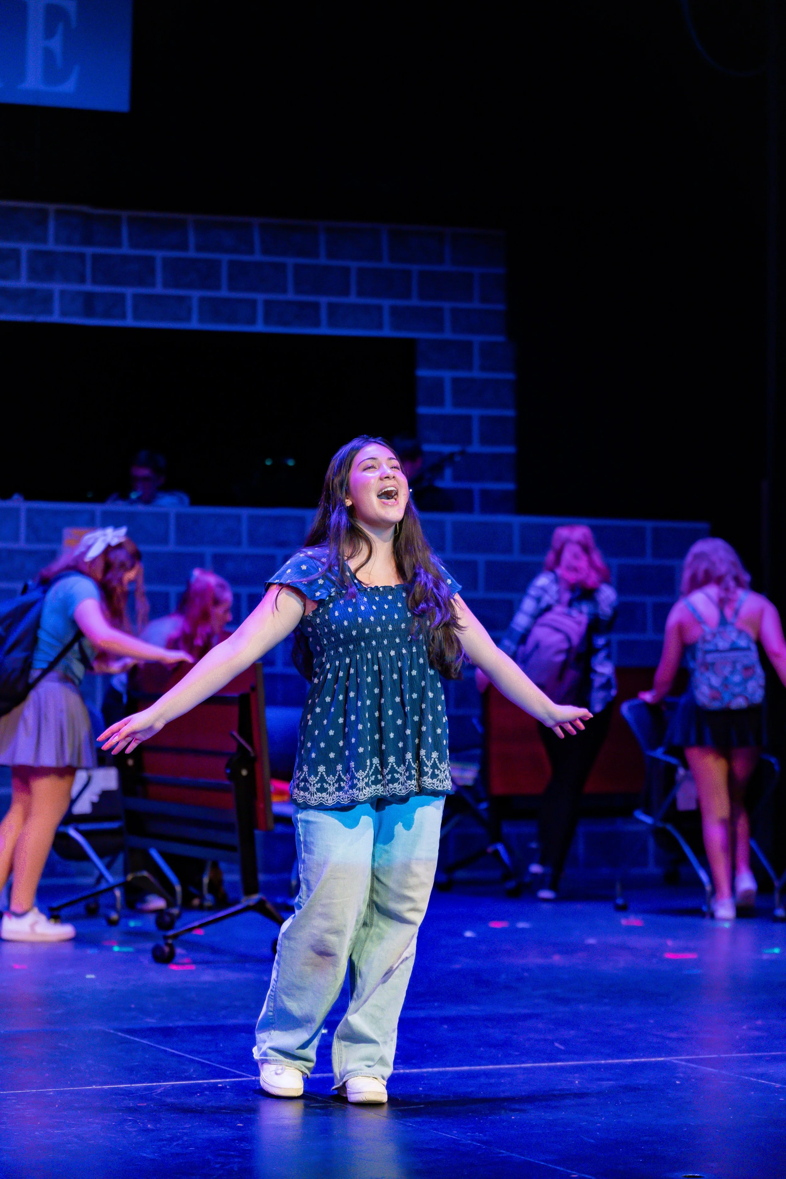 A young woman in a blue polka dot shirt and light-colored pants performing on stage with arms outstretched and a joyful expression, surrounded by other performers in casual clothing under blue stage lighting.
