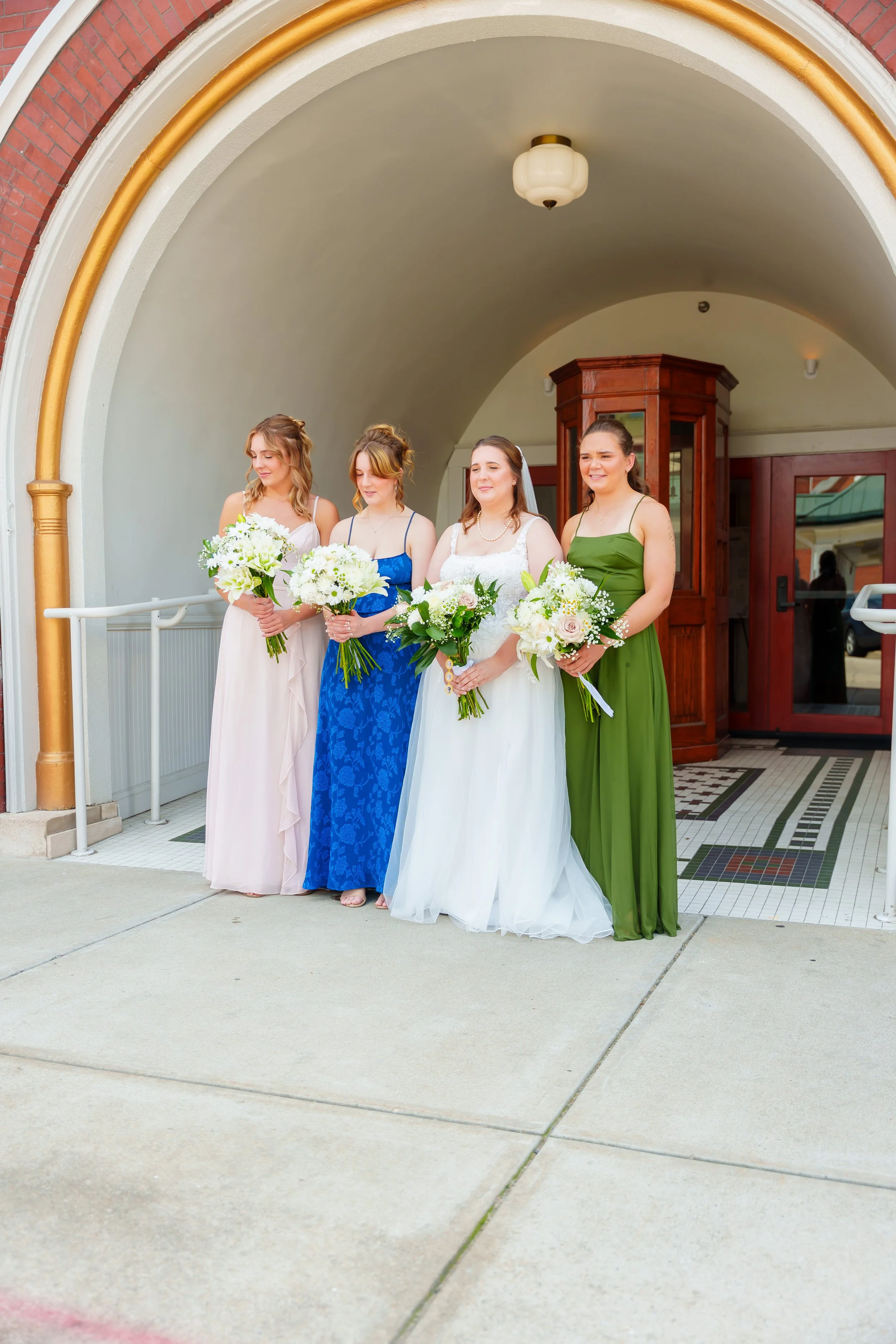 Four women in formal dresses holding bouquets stand outside under an arched entrance with a red brick and cream-colored wall, a wooden door, and a tiled floor.